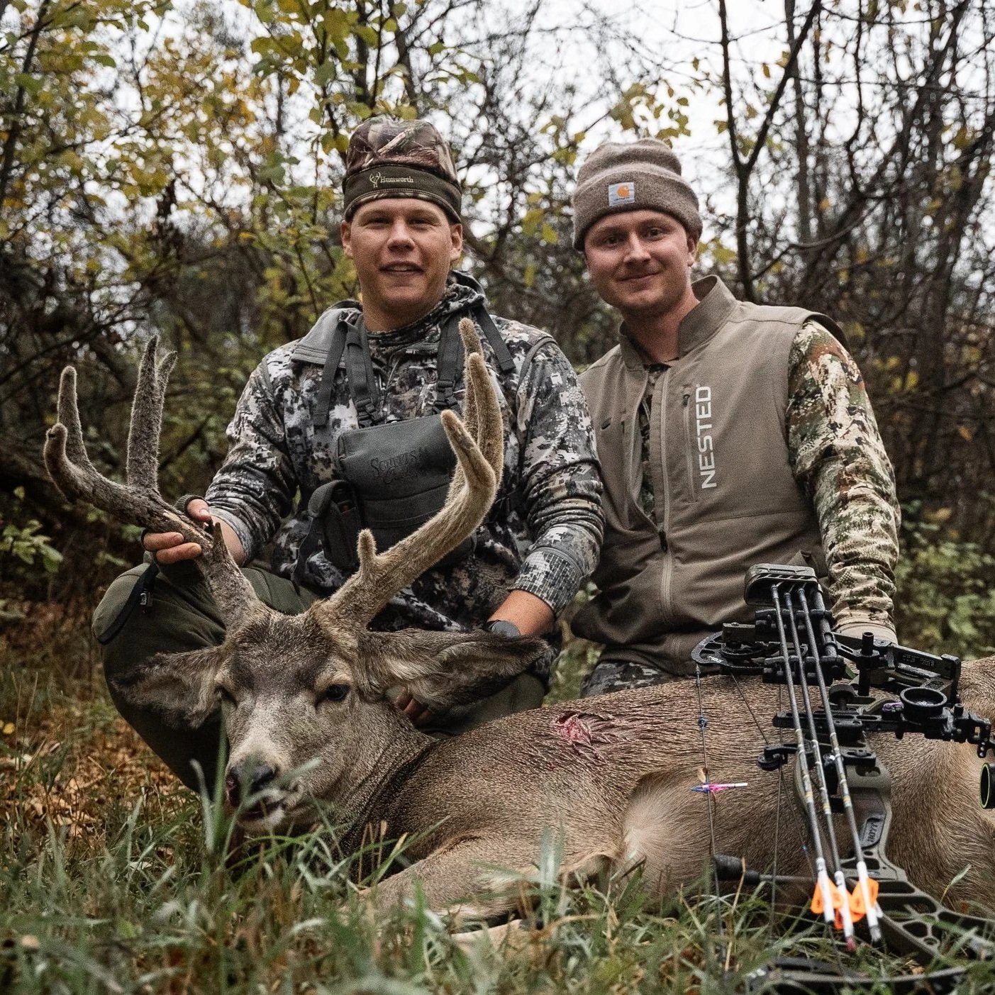 Two hunters posing with a recently hunted deer in a forest setting, holding the deer's antlers, with a crossbow lying on the ground beside them.