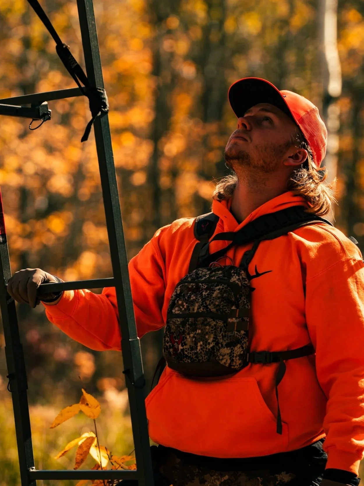 A man with long hair, wearing an orange hoodie and a red cap, stands outdoors with a fall forest background. He is looking upward while holding onto a metal structure.