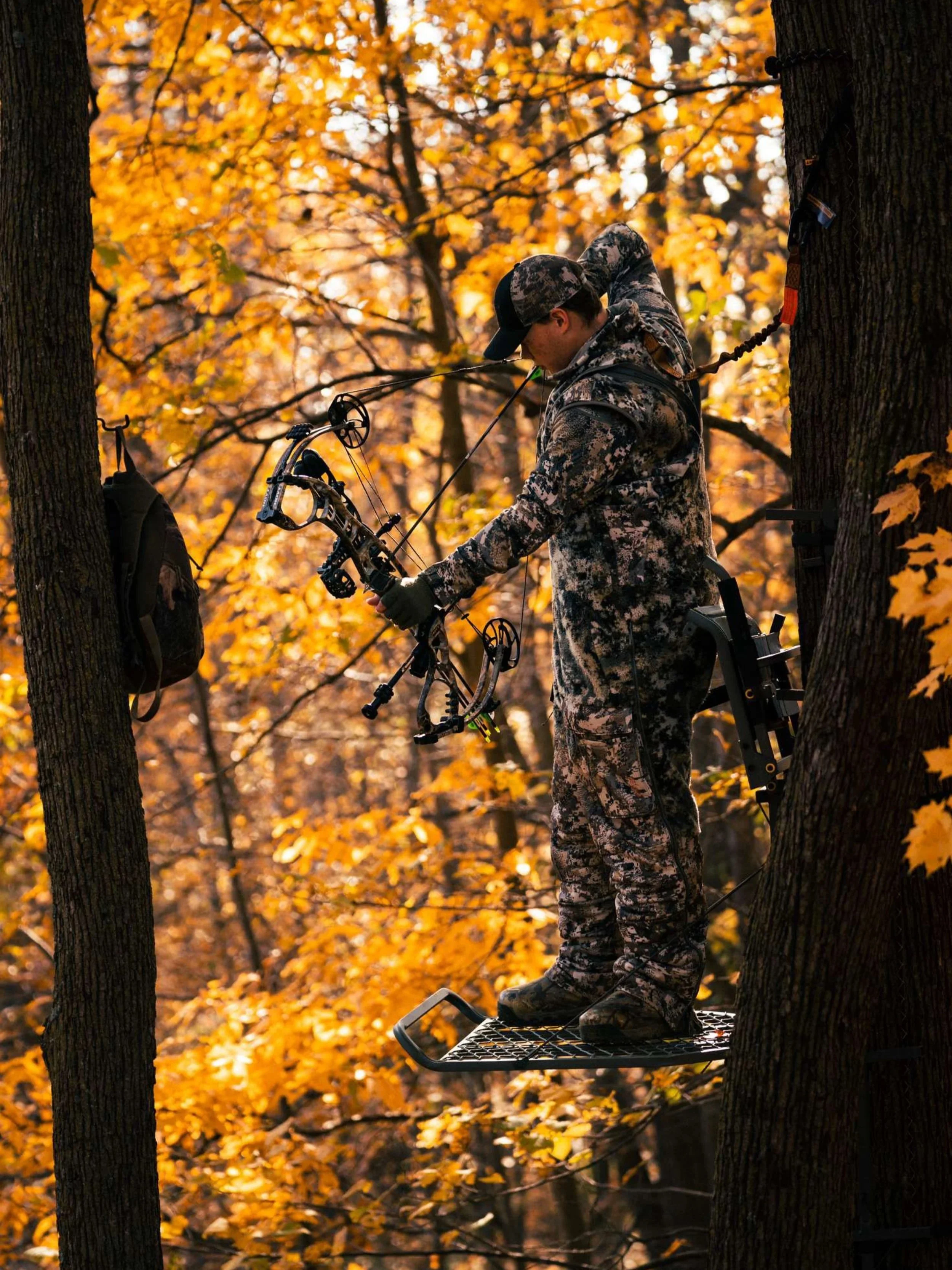 A person dressed in camouflage clothing stands on a platform between two trees in a forest with orange and yellow fall leaves, preparing to shoot a bow and arrow.