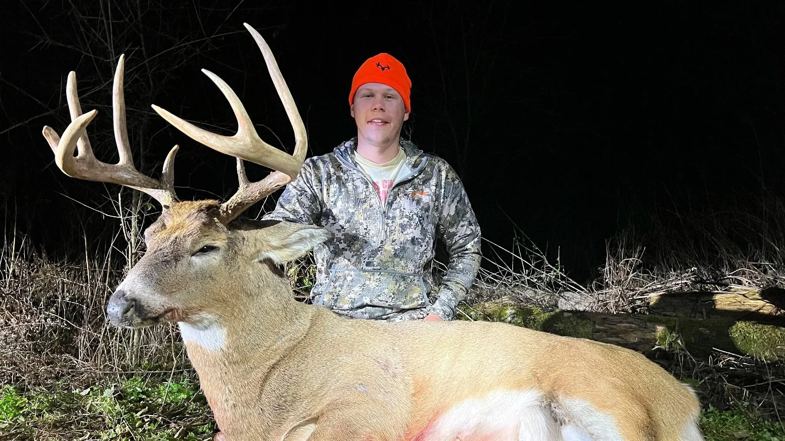 A young man wearing a bright orange beanie and camouflage clothing kneels next to a large deer with antlers, in a wooded outdoor area at night.