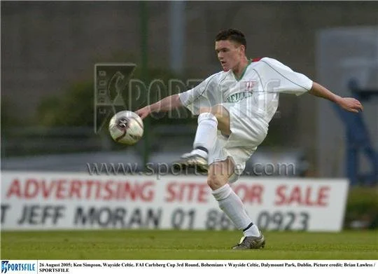 A soccer player in a white uniform kicking a ball on a field with advertising banners in the background.