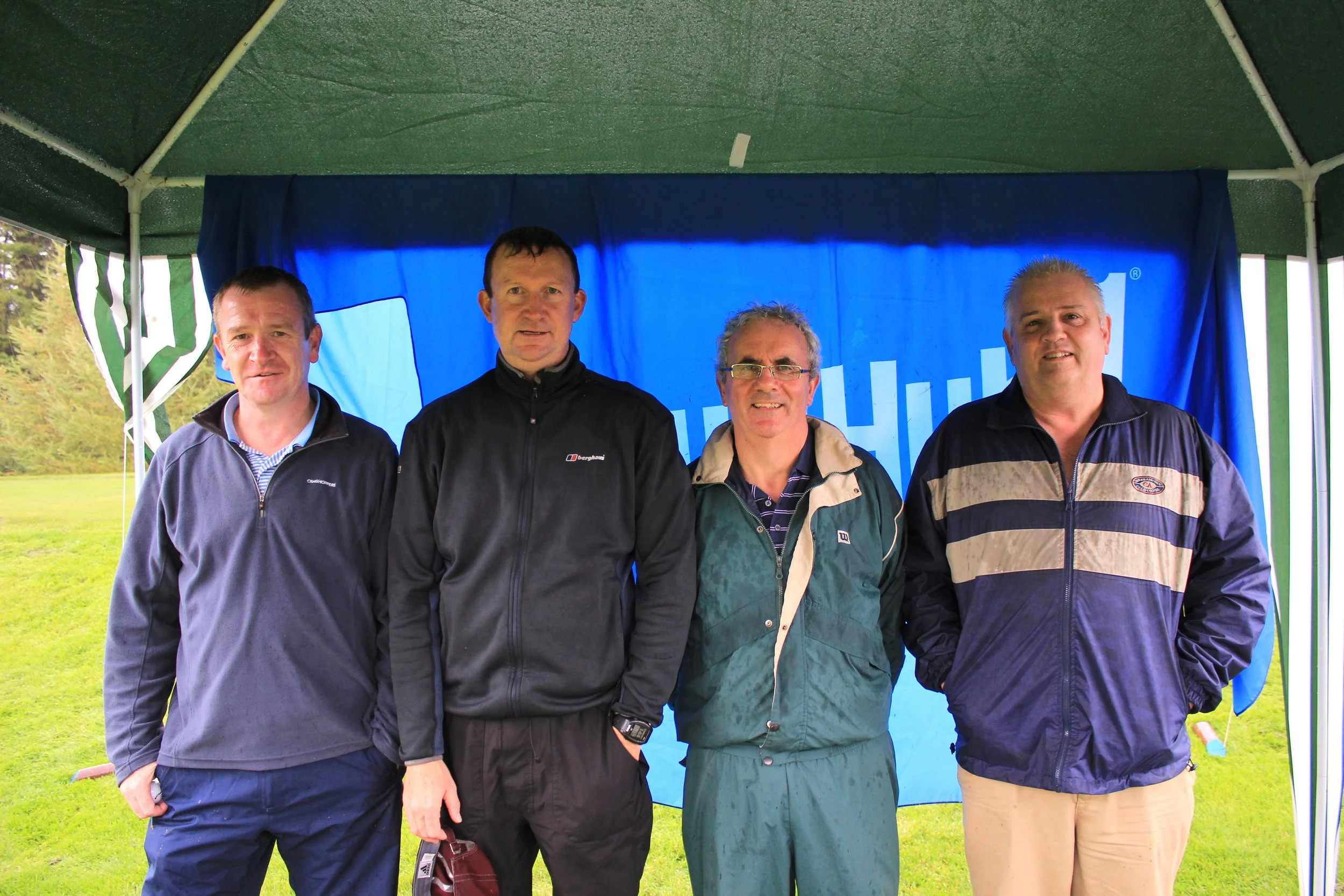 Four men standing under a green tent with a blue banner behind them. The men are dressed in casual jackets and shirts, smiling at the camera outdoors.