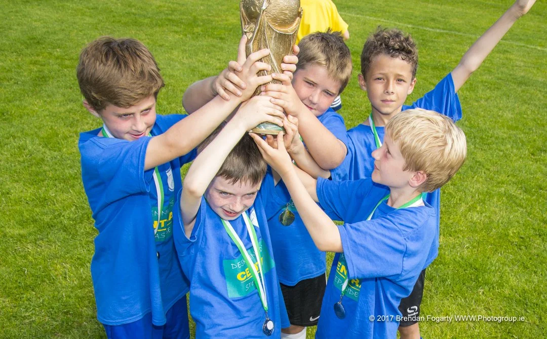Young boys in blue sports jerseys celebrating and lifting a trophy on a grassy field after winning a game