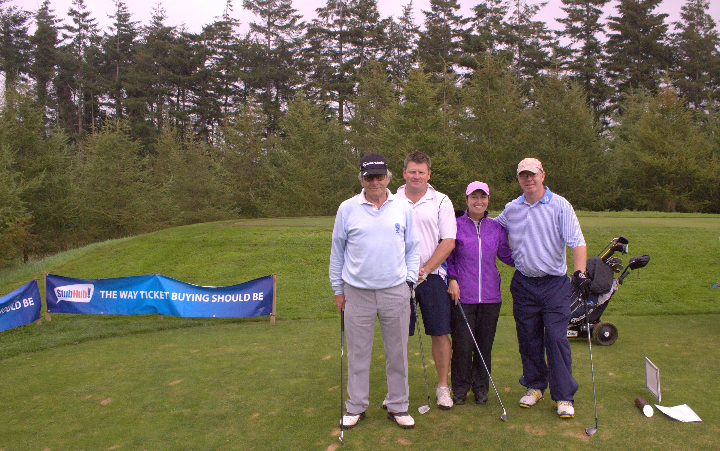 Four people standing on a golf course with trees in the background, posing for a photo. Three men and one woman, all dressed in golf attire, holding golf clubs. A golf bag with clubs is visible to the right. A banner with the text 'StubHub! THE WAY T