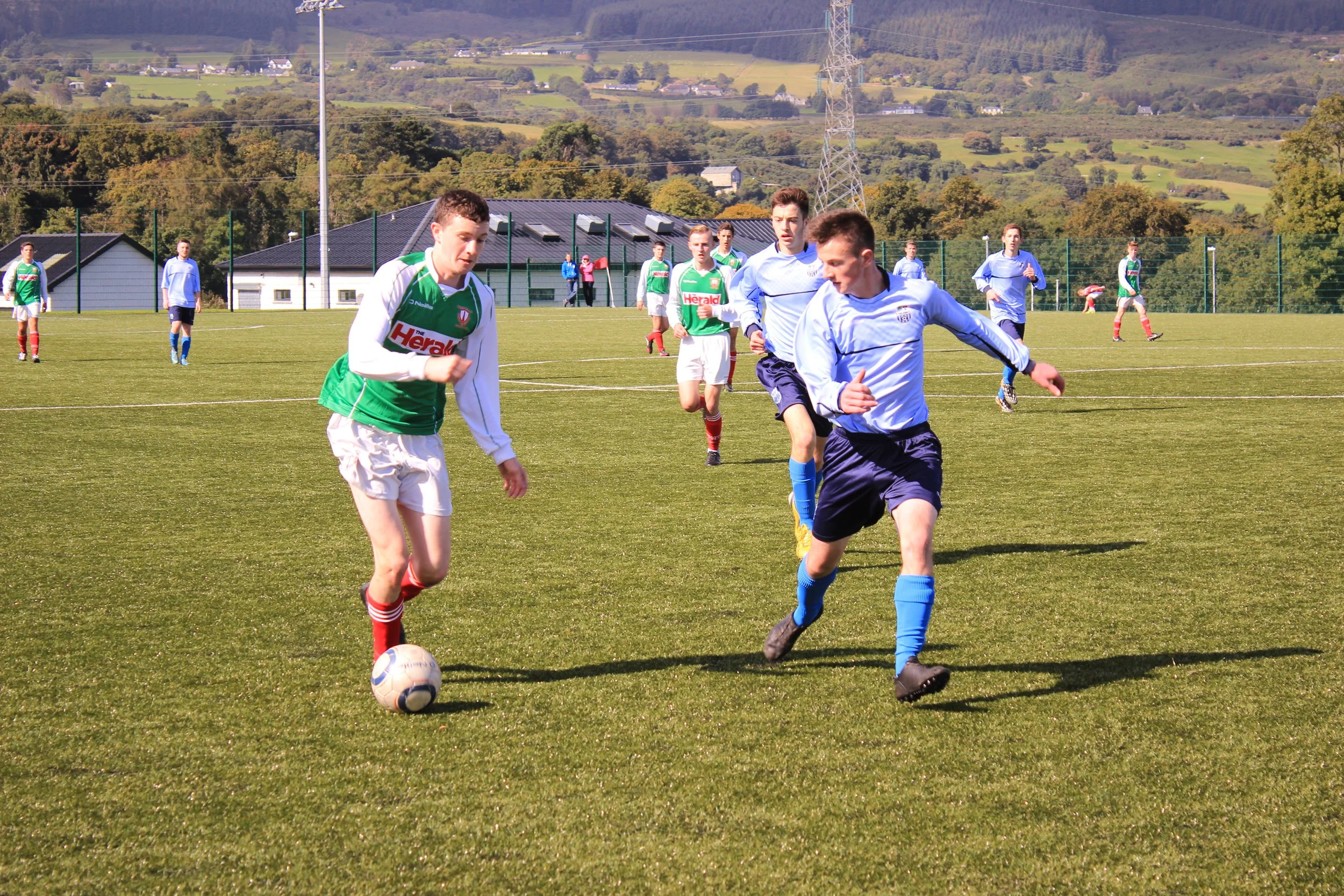 A group of young men playing soccer on a grassy field, with some wearing white jerseys and others in green jerseys, and a scenic background of trees, hills, and buildings.