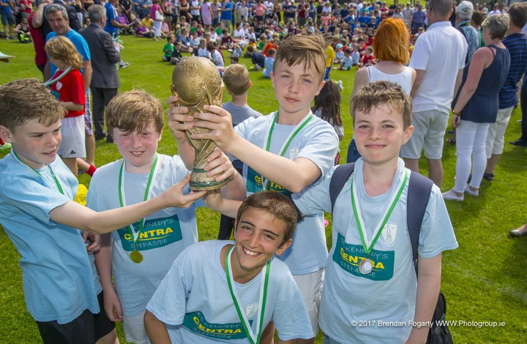Group of young boys in light blue shirts holding a golden trophy, celebrating a sports victory outdoors with a large crowd in the background.