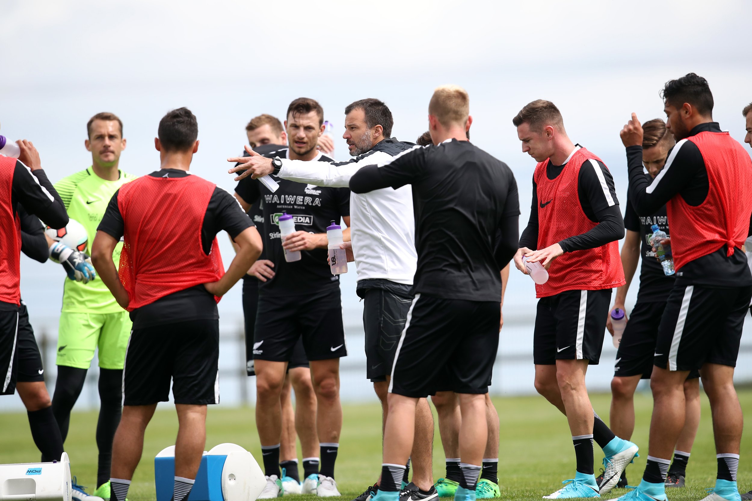 Soccer coach giving instructions to players during a practice session on a grassy field. The players are wearing black and red sports gear, and some are holding water bottles.