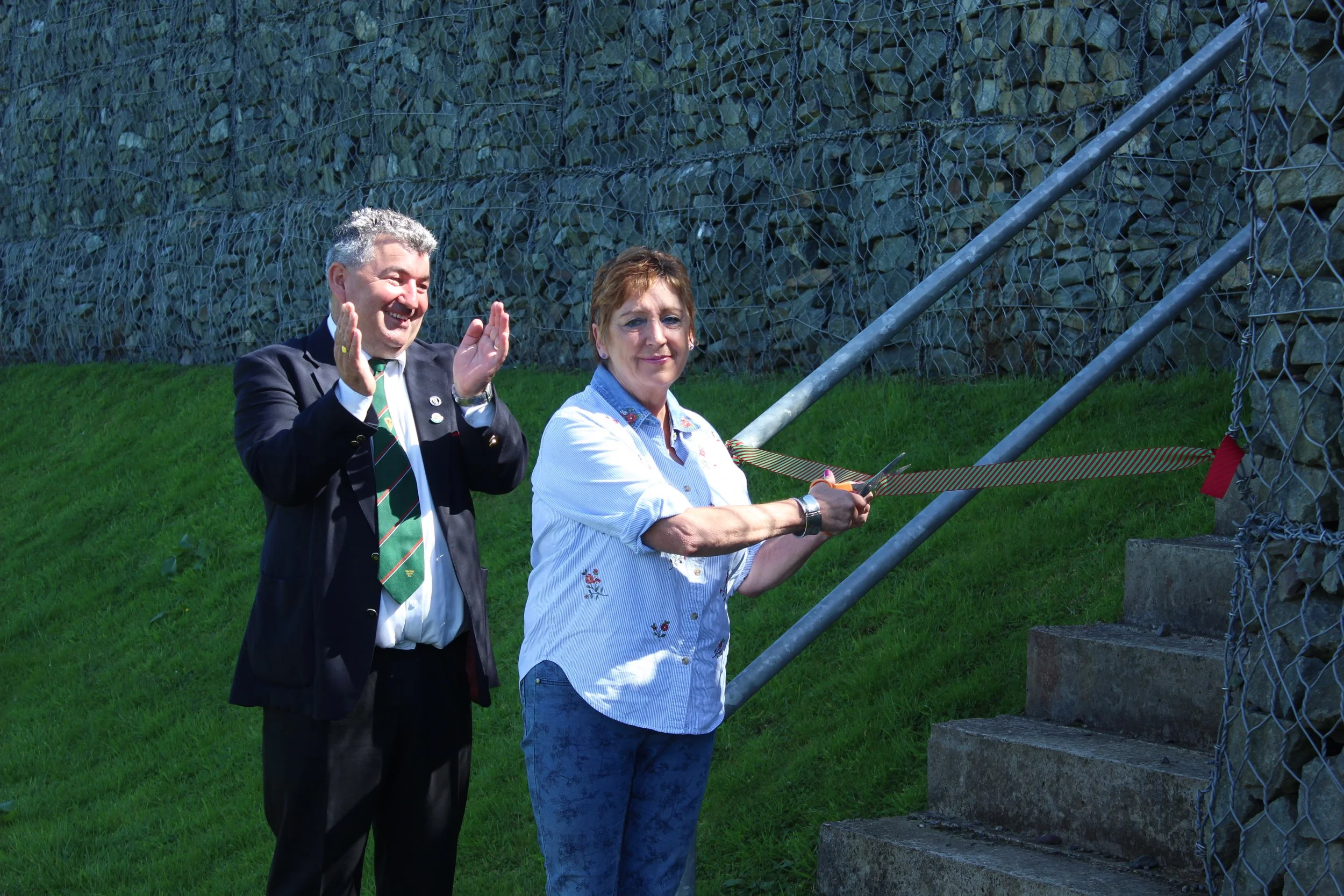 A man in a suit and a woman in a light blue shirt cut a ribbon with scissors at an outdoor event, standing in front of a gravel wall and a chain-link fence.
