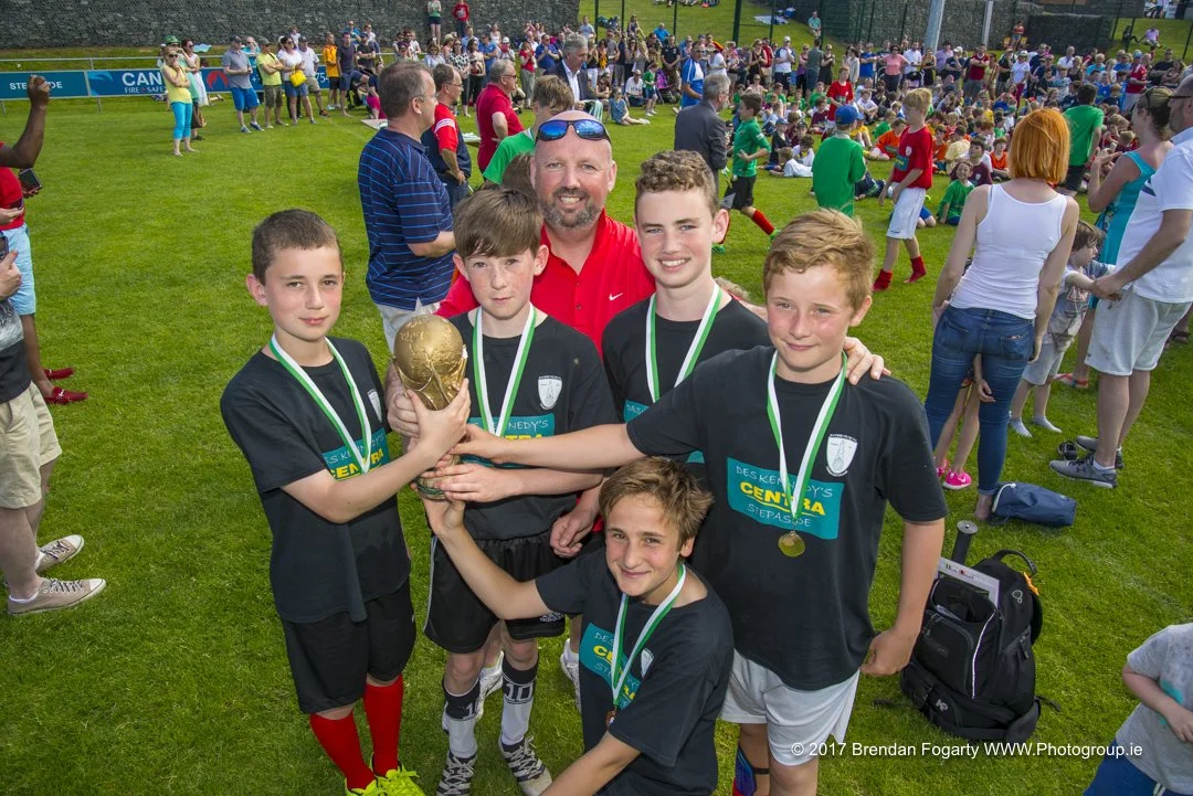 Group of young boys and coach celebrating with a trophy after a soccer game, on a grassy field filled with spectators.
