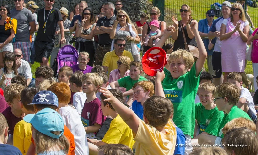 Children and adults gathered outdoors, some children sitting on the ground and raising their hands, while some adults stand clapping and watching a children's event on a sunny day.