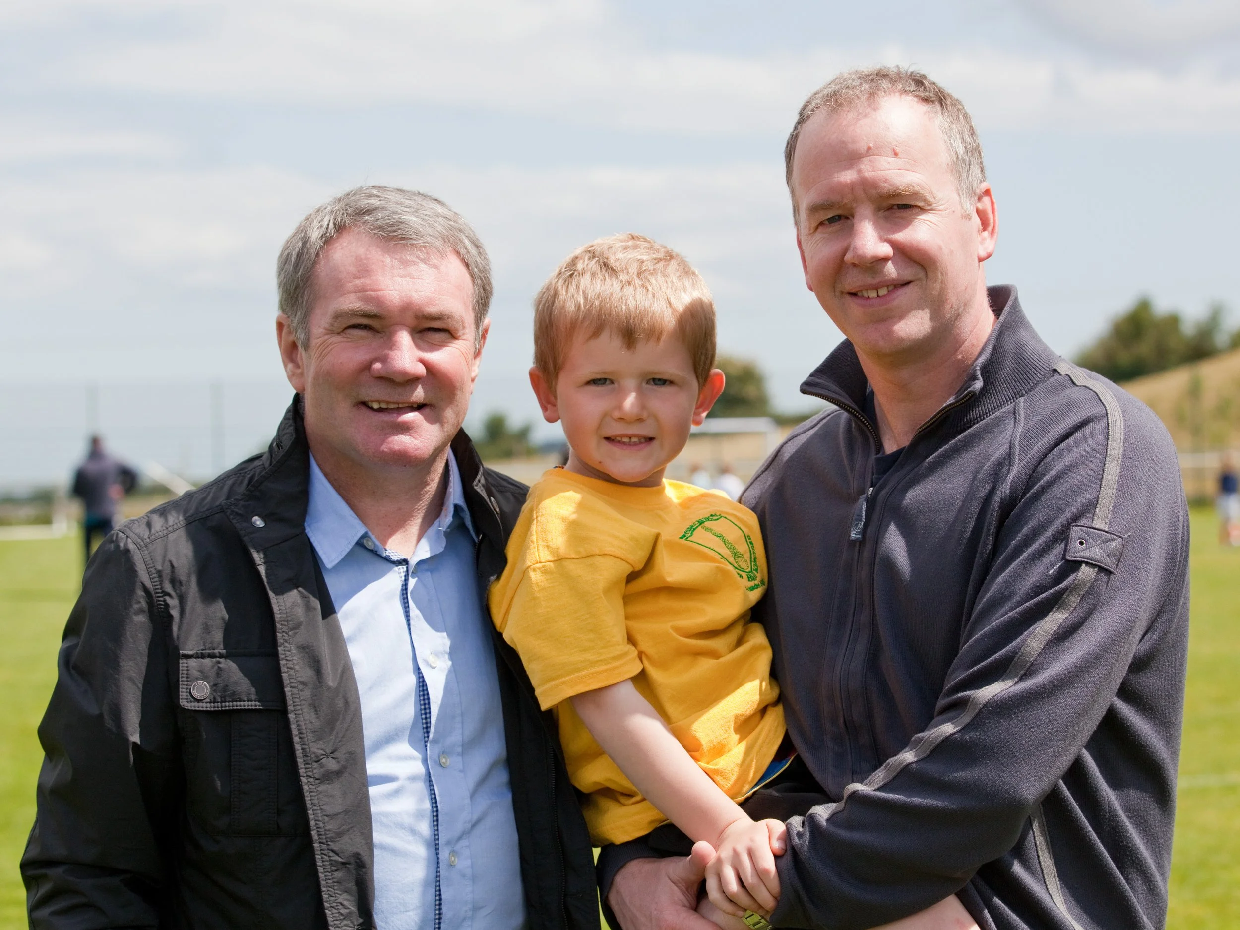 Three males and a boy smiling outdoors with a grassy field and blue sky in the background.