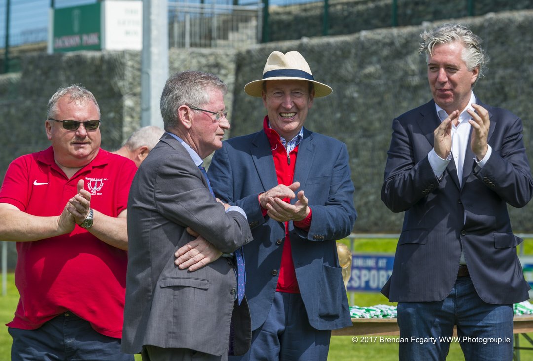 Four men standing outdoors, dressed in business and casual attire, clapping and smiling during a sunny day, with stadium or sports field in the background.