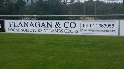 A large outdoor sign advertising Flanagan & Co, local solicitors at Lambs Cross, on a grassy area with a tennis court in the background.