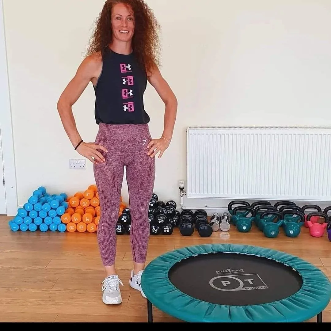 Woman with curly red hair standing in a gym, wearing athletic leggings, a sleeveless top, and sneakers, smiling with hands on hips. Behind her are stacks of colorful dumbbells, kettlebells, and a small trampoline.