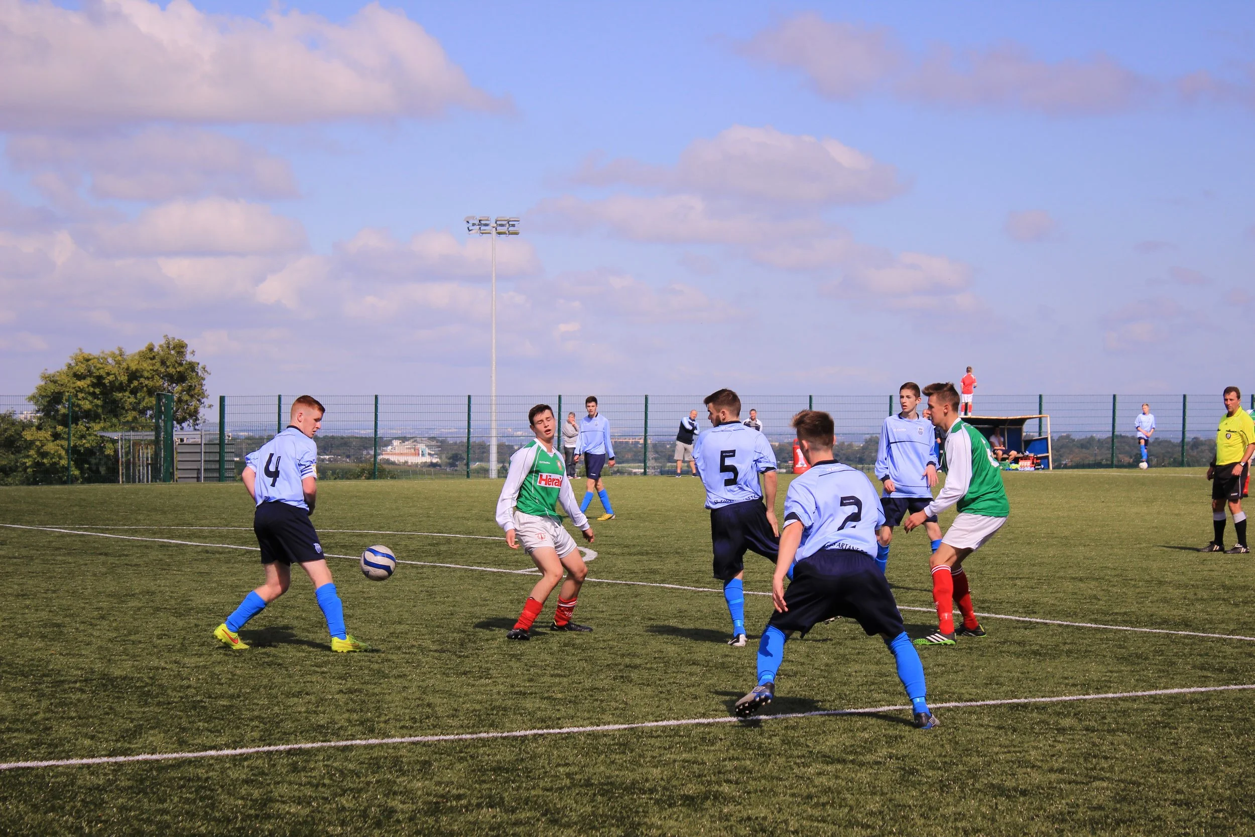 Soccer players on the field during a match under a blue sky with clouds, with some players in blue uniforms and others in green and white uniforms.