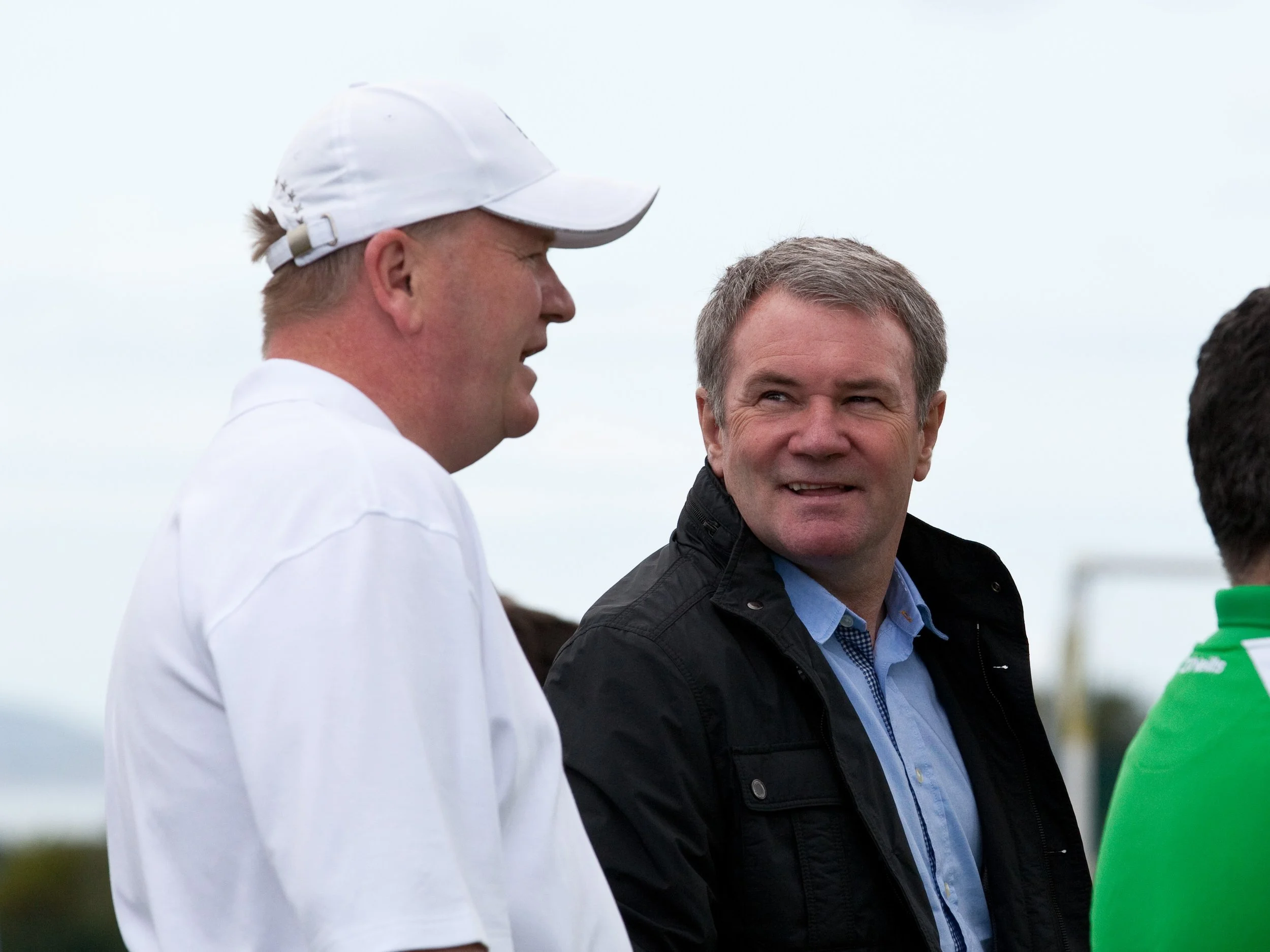 Three men are engaged in conversation outdoors on a cloudy day. One person wears a white cap and shirt, another has light brown hair and wears a black jacket over a blue shirt, and the third, partly visible, wears a green shirt.