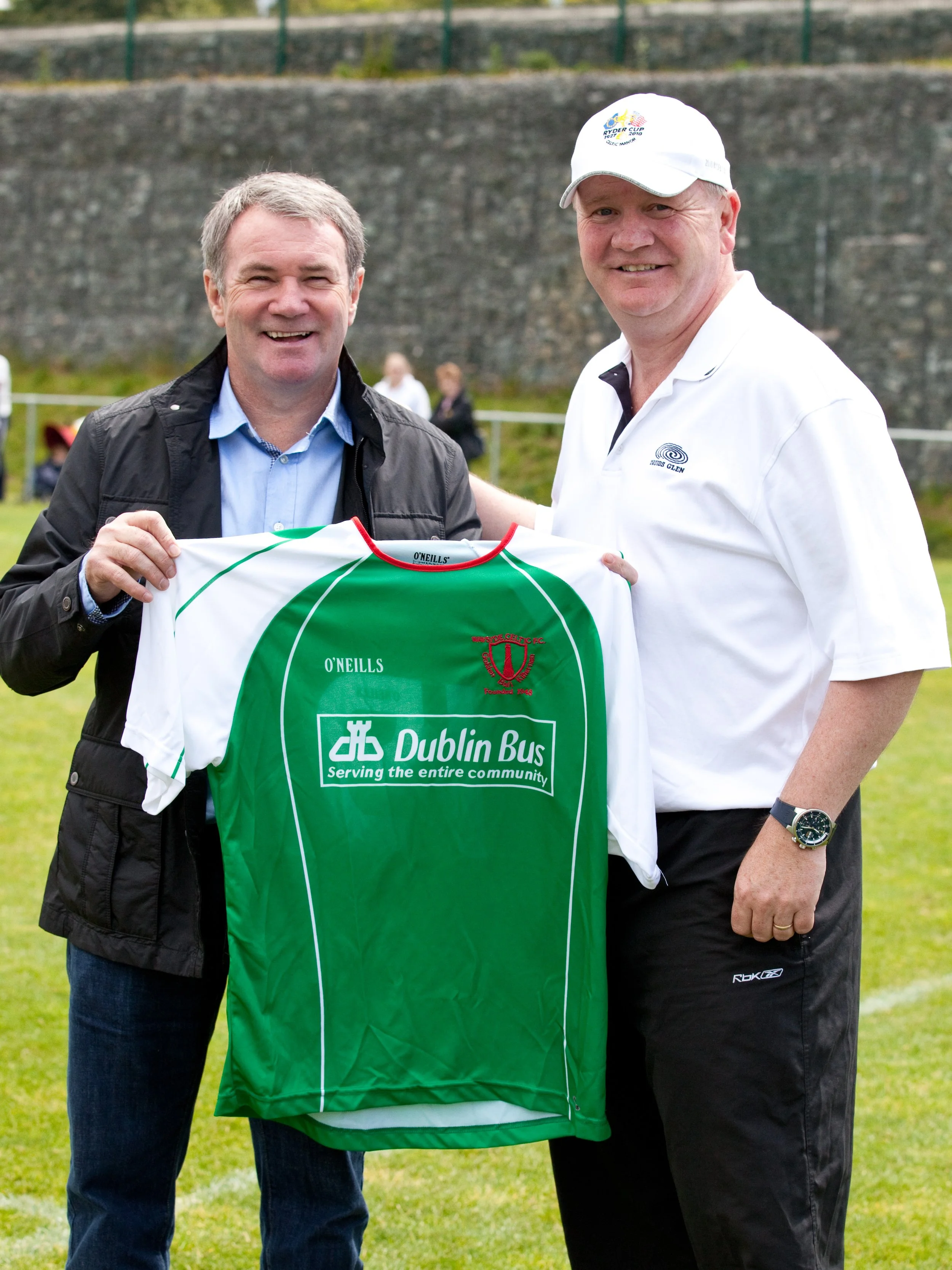 Two men holding a green and white sports jersey at an outdoor event, with a stone wall and green grass in the background.