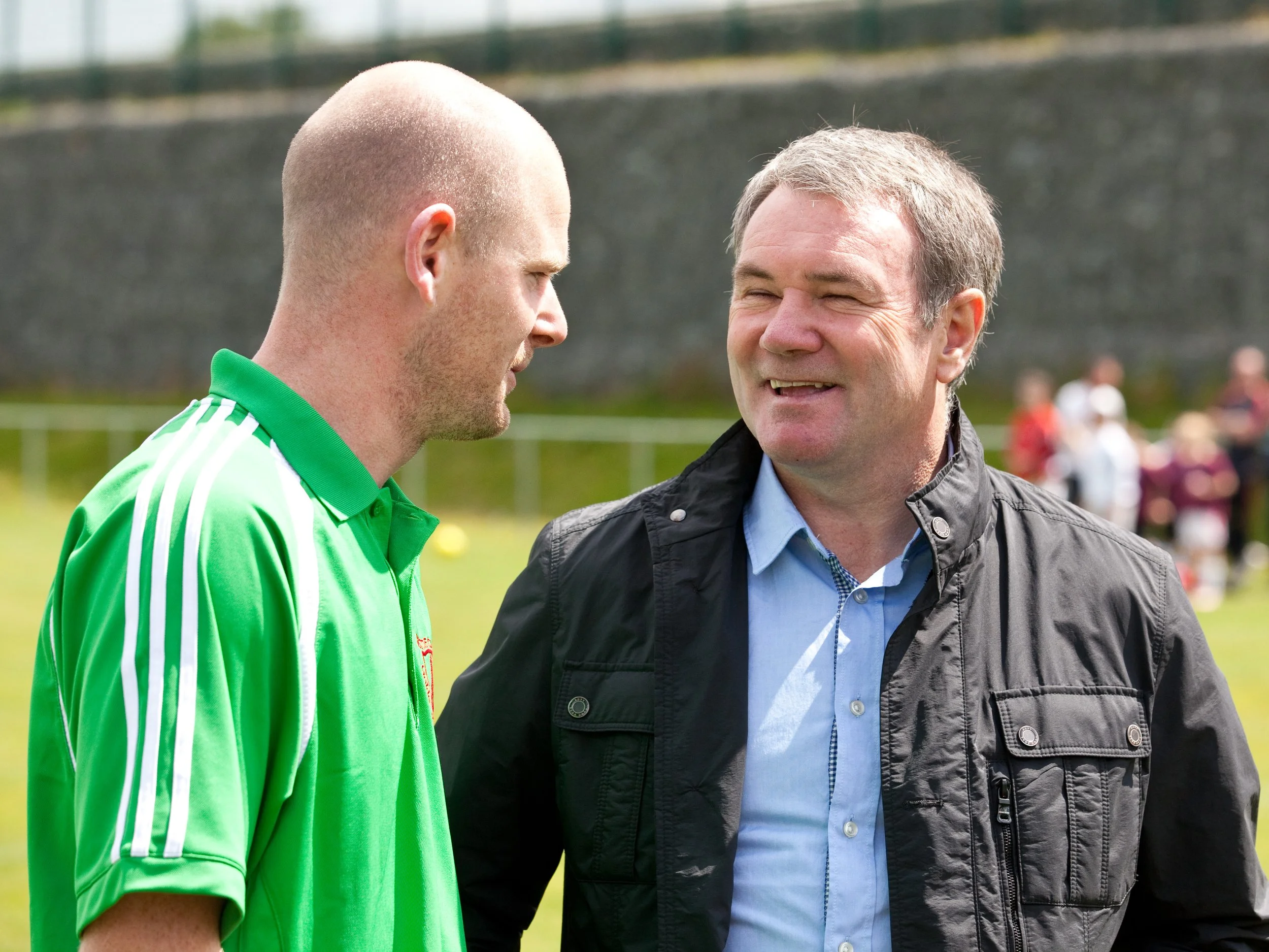 Two men are standing outdoors on a soccer field, facing each other and smiling. The man on the left is wearing a bright green sports jersey and has a shaved head, while the man on the right is wearing a black jacket over a light blue shirt and has sh