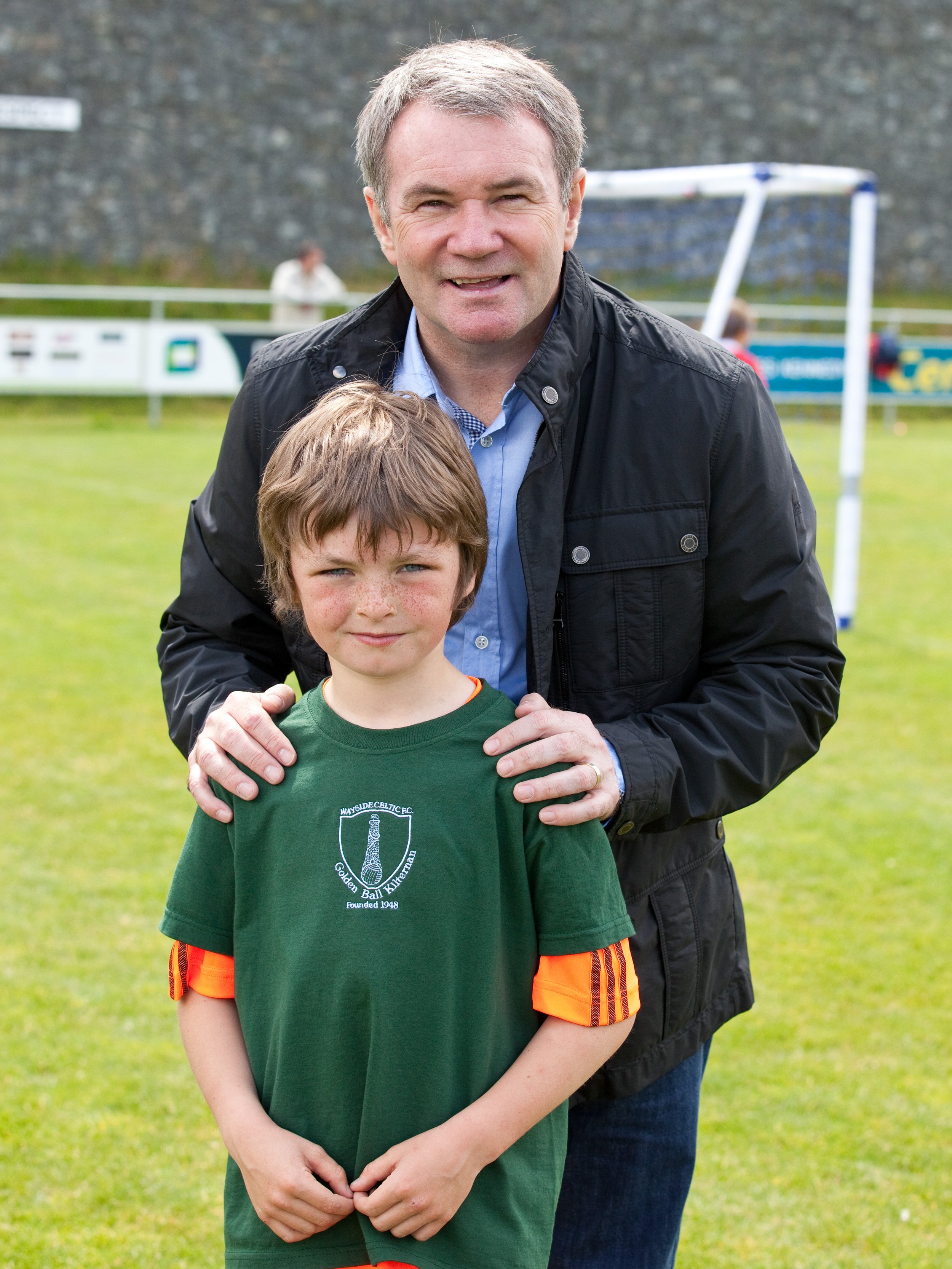 A middle-aged man with gray hair smiling, standing behind a young boy with a serious expression, on a grassy sports field with soccer goals in the background.