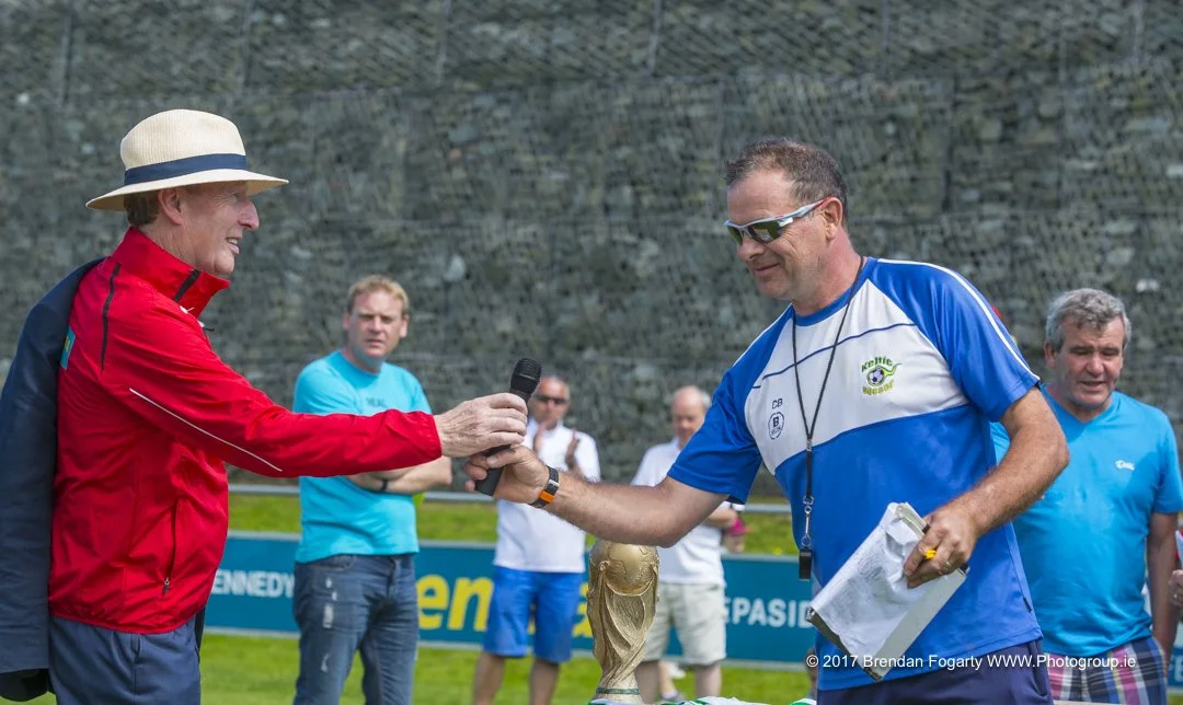 A woman in a red jacket and hat presenting a trophy to a man in a blue sports shirt at an outdoor event with several onlookers.