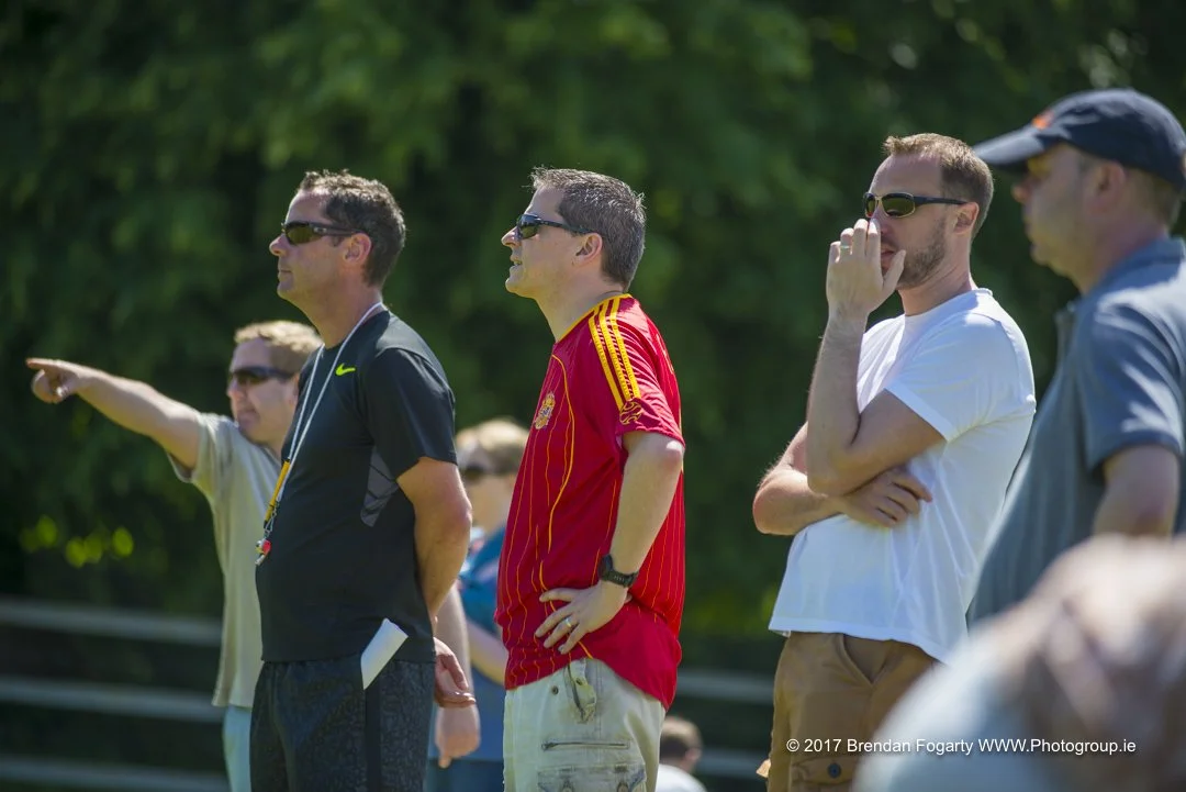 Group of men watching a sports game outdoors, some wearing sunglasses, one pointing, one with a whistle, and others with hand on chin, in sunny weather with green trees in the background.