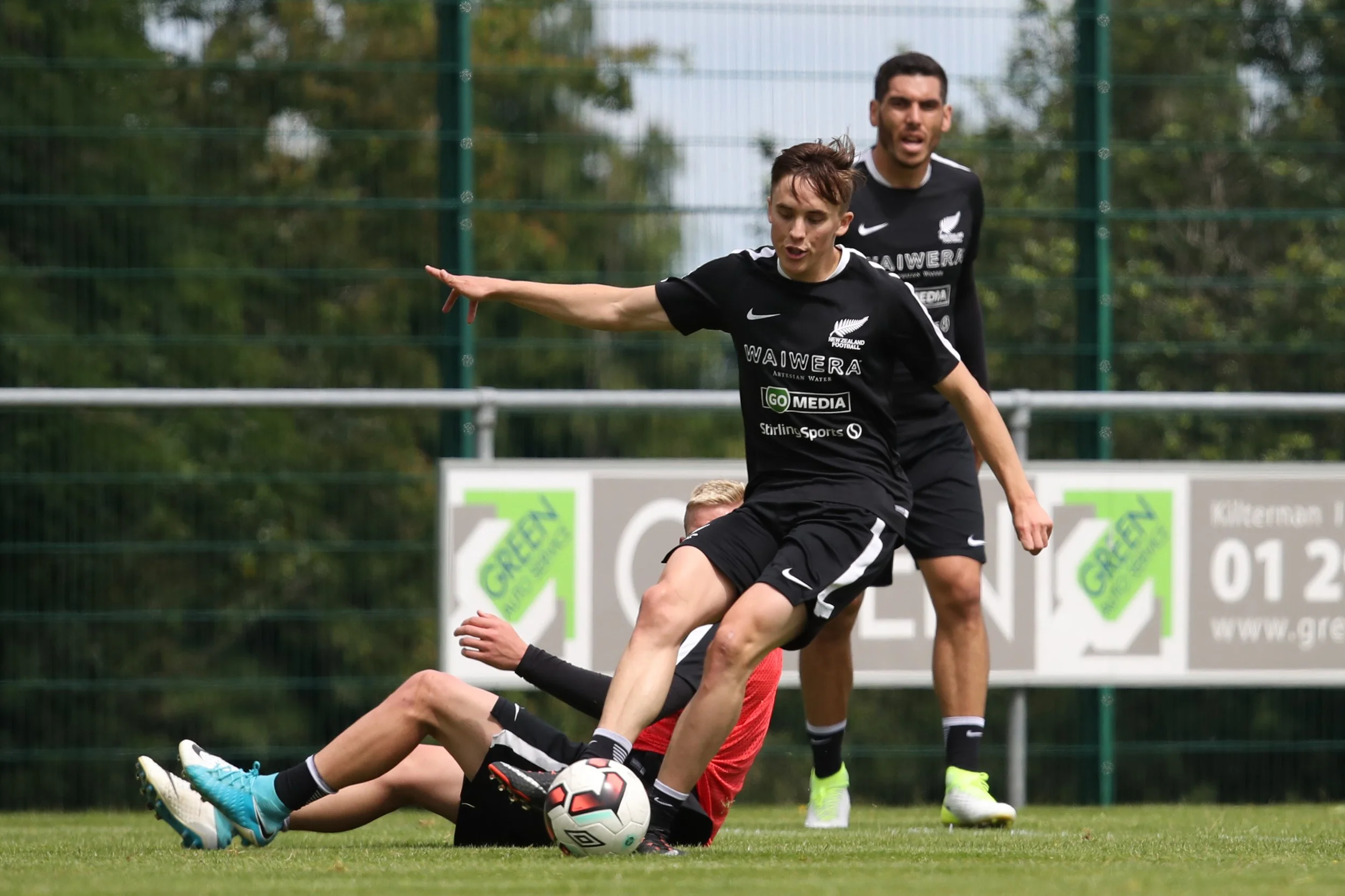 Two soccer players in black jerseys compete for possession of a soccer ball during a game on a grassy field, with a green fence and trees in the background.