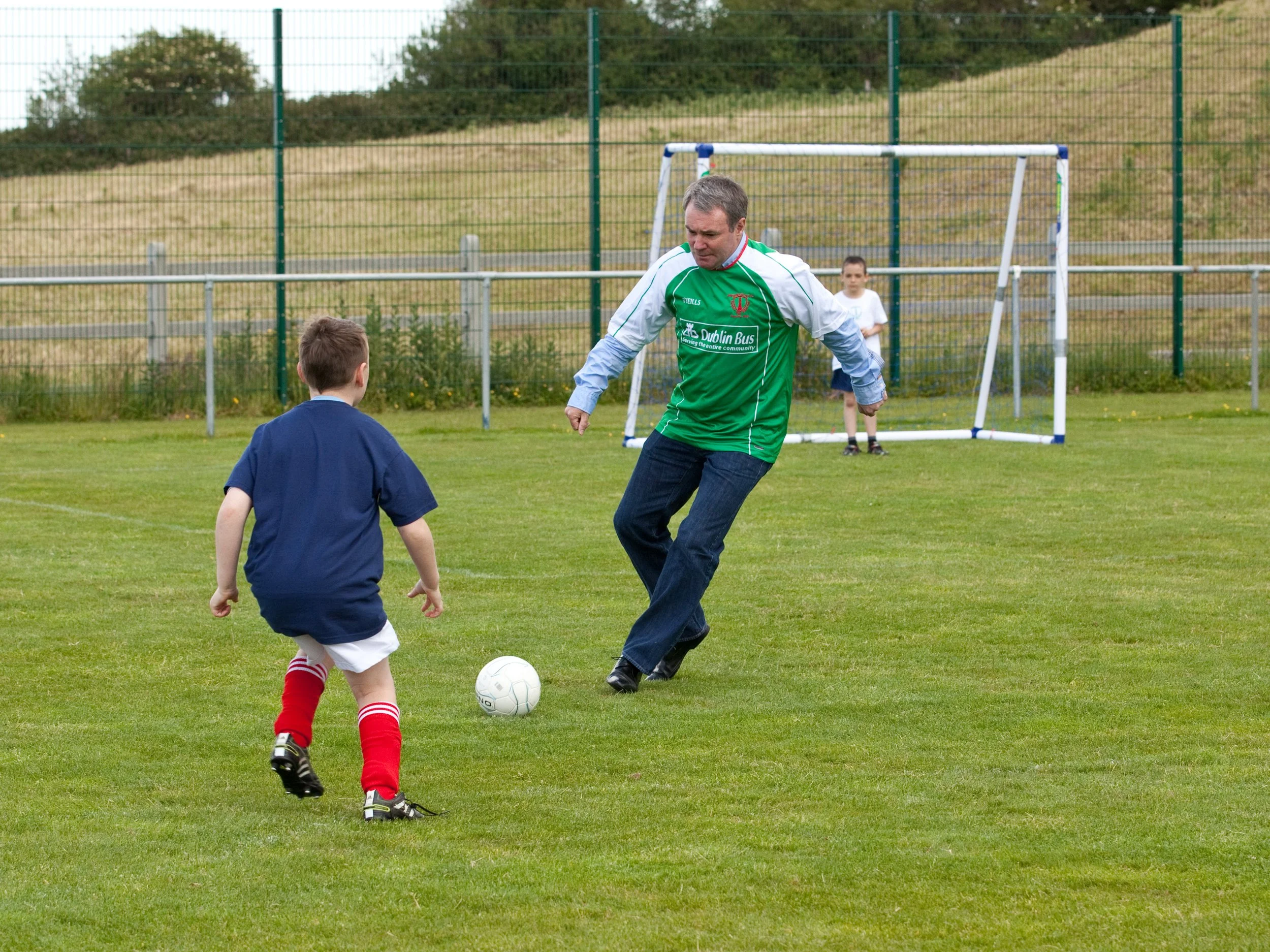 A man in a green and white sports jersey playing soccer with two children on a grassy field, with a soccer goal in the background.