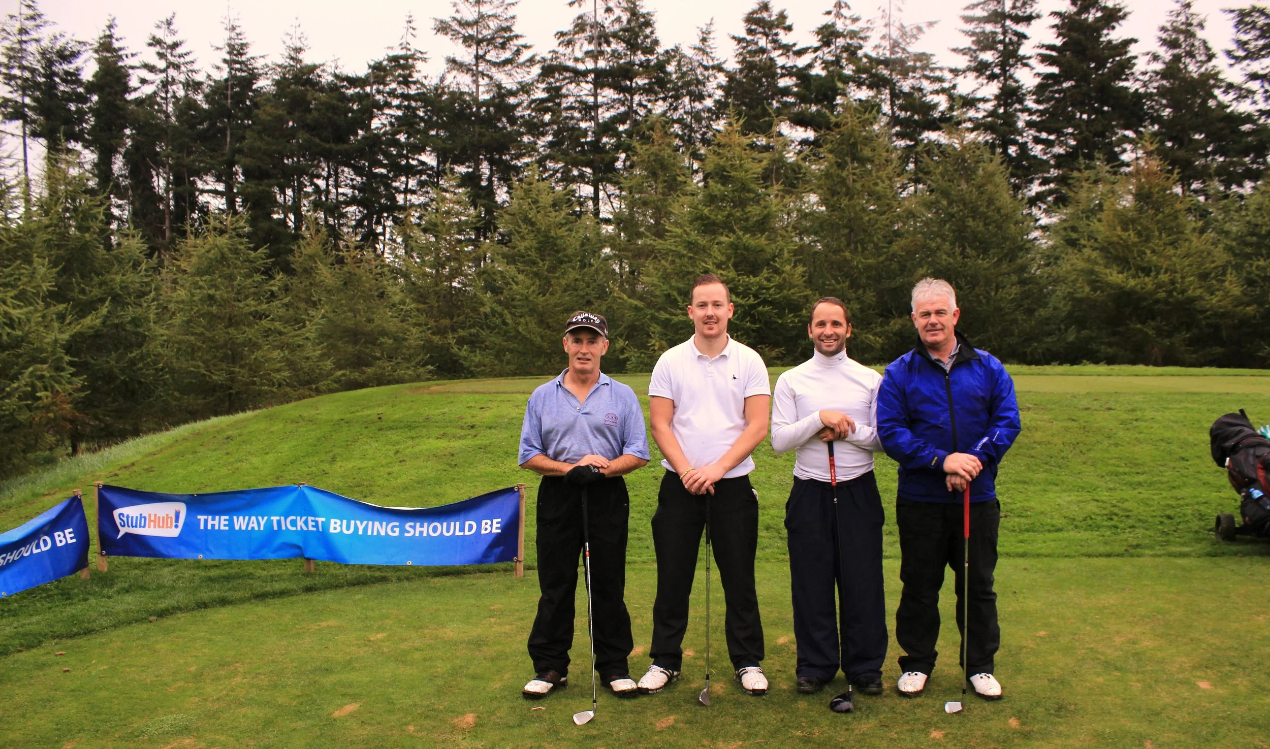 Four men with golf clubs standing in front of a blue banner at a golf course. Trees in the background. The banner reads "StubHub! THE WAY TICKET BUYING SHOULD BE."