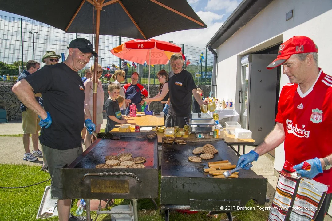 People grilling hamburgers and hot dogs at an outdoor event with tables, umbrellas, and spectators in the background.