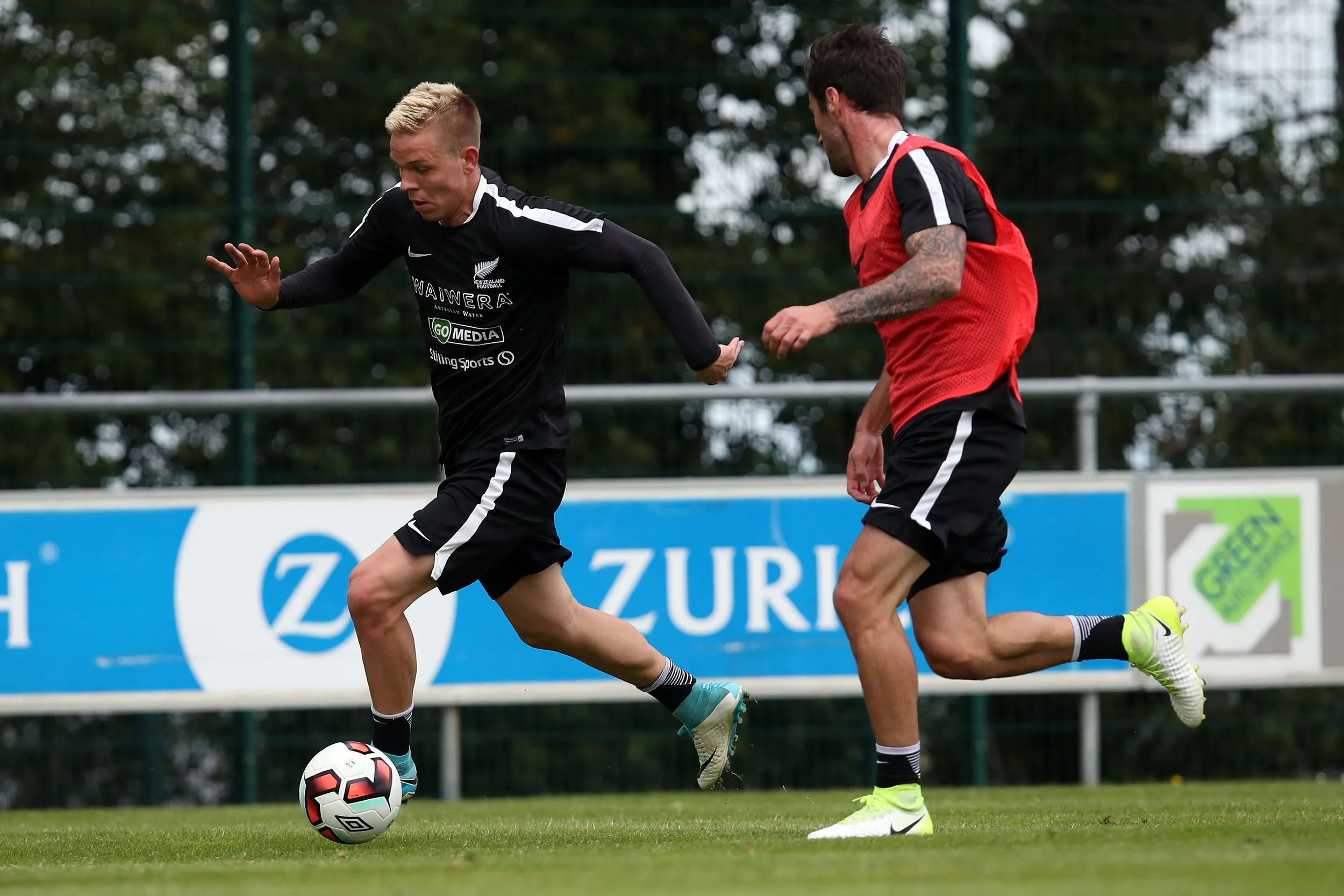 Two male soccer players are competing for the ball during a match on a green grass field, with one in a black uniform and the other in a red vest, both wearing cleats.