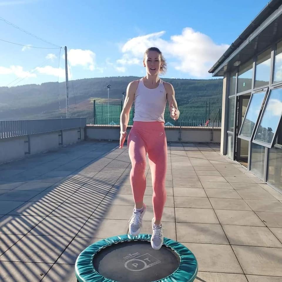 A woman jumping on a small trampoline on a rooftop terrace with a mountain landscape in the background.