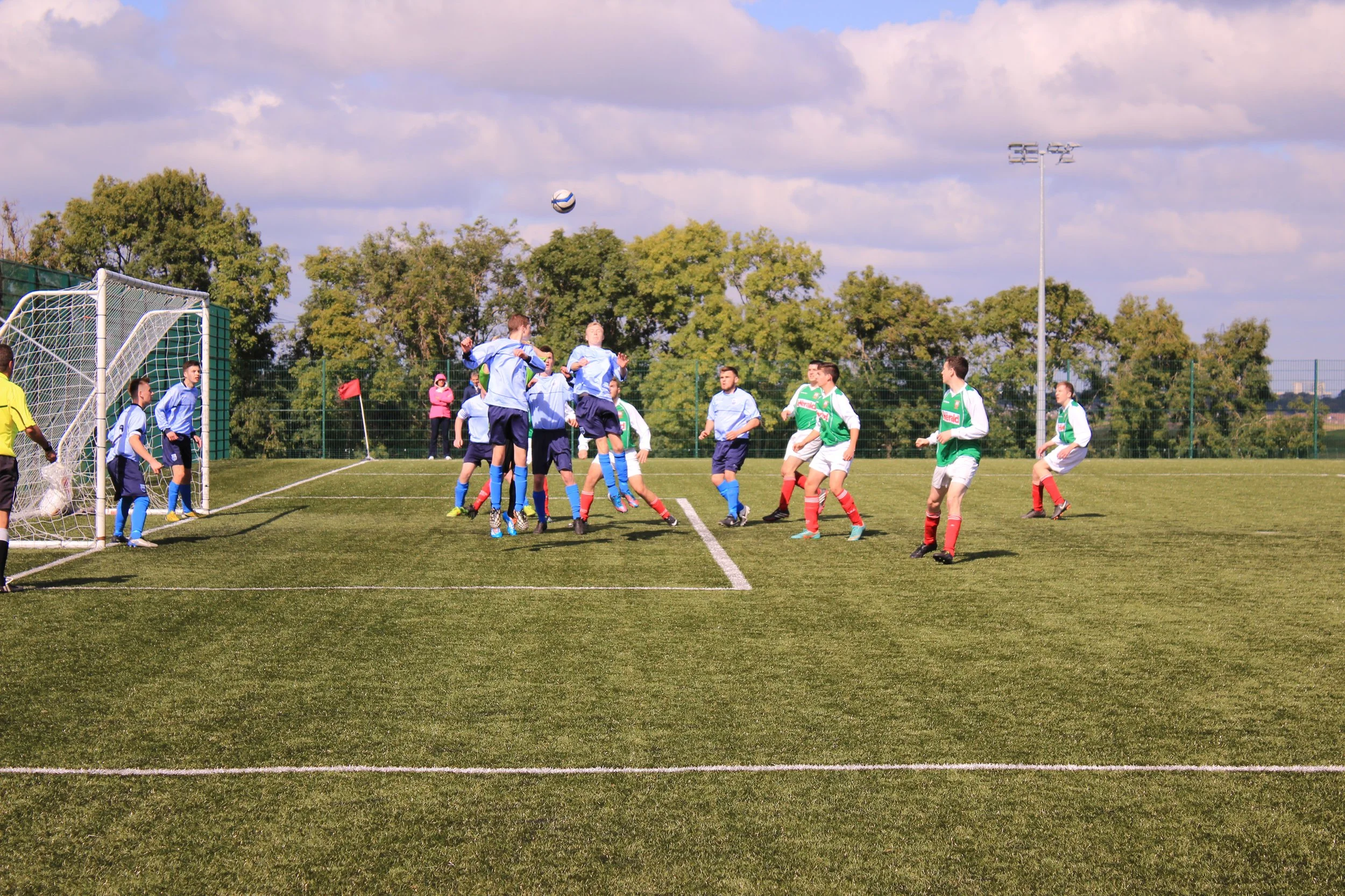 Young soccer players in light blue and green uniforms compete for a soccer ball during a match on a grassy field, with trees and cloudy sky in the background.