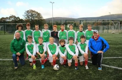 A youth soccer team with 11 boys and 2 coaches posing on a soccer field, wearing green and white uniforms.