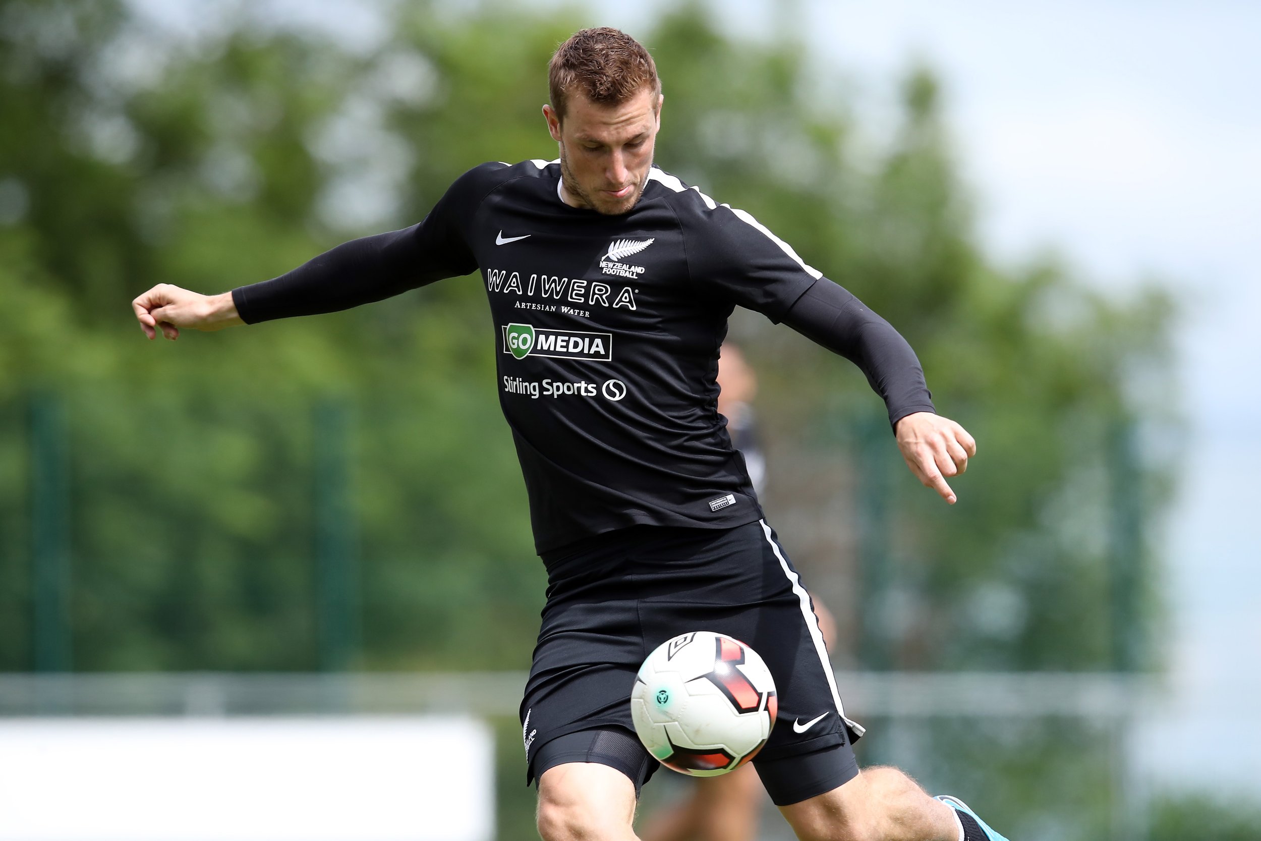 A male soccer player wearing a black jersey with sponsors' logos, playing with a soccer ball on an outdoor field with green trees in the background.