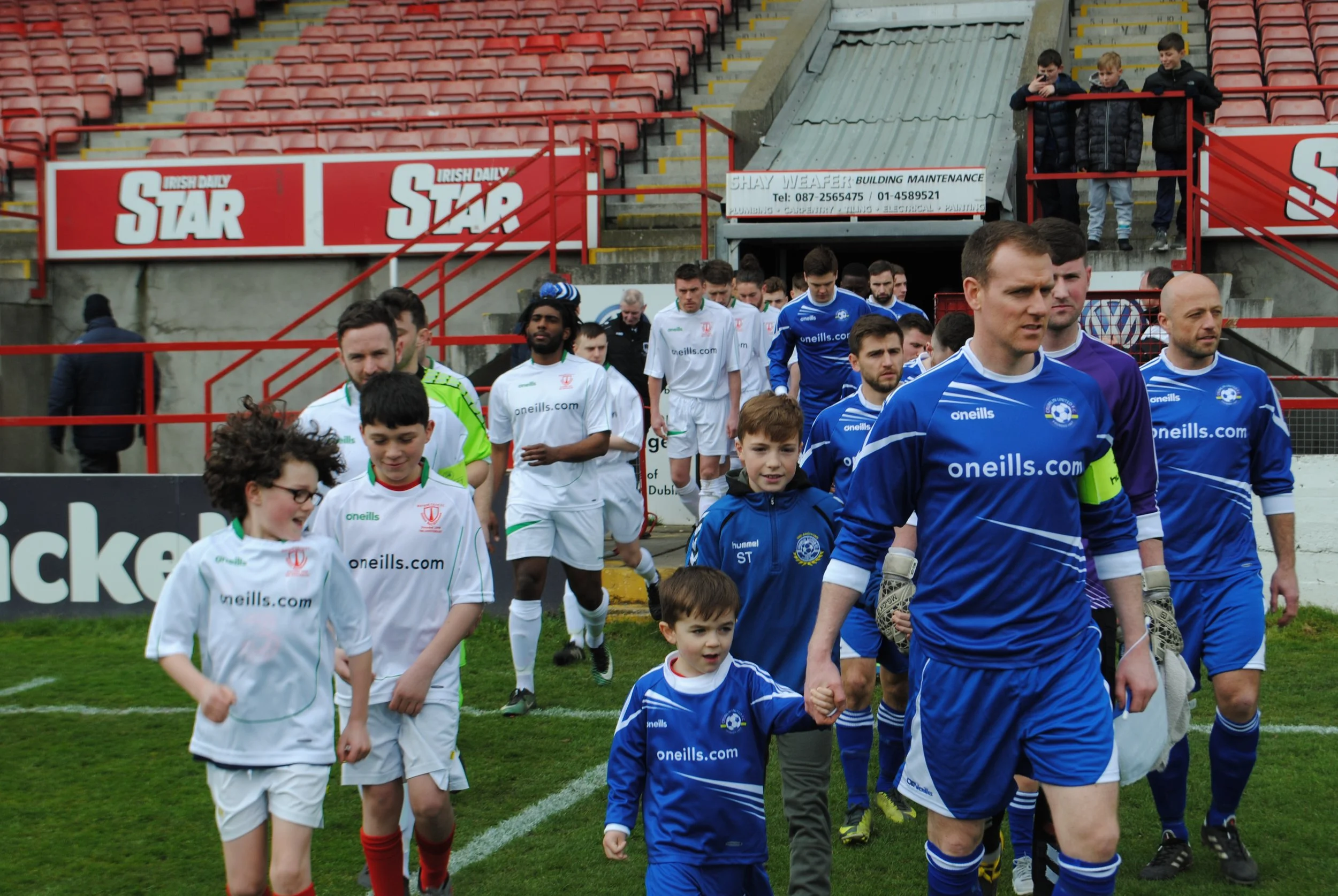 Soccer players walking onto the field before a game, with children and adults in the background at a stadium with red seating.
