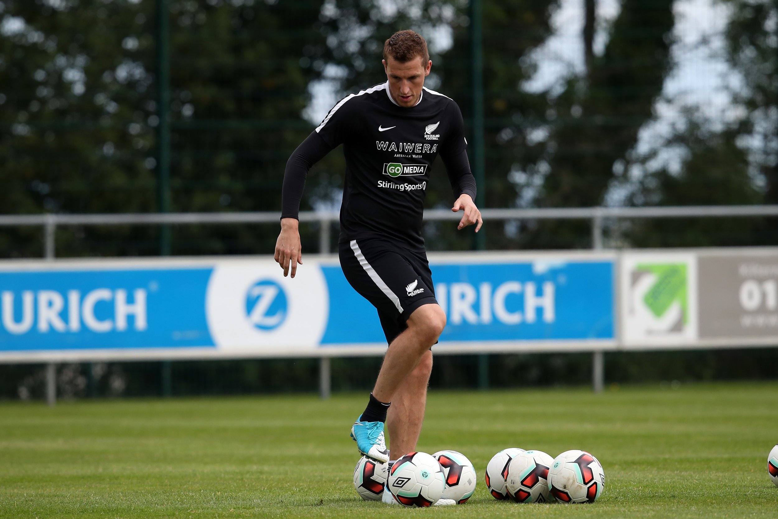 A male soccer player on a field practicing with multiple soccer balls, wearing a black jersey with sponsorship logos, black shorts, and blue shoes.