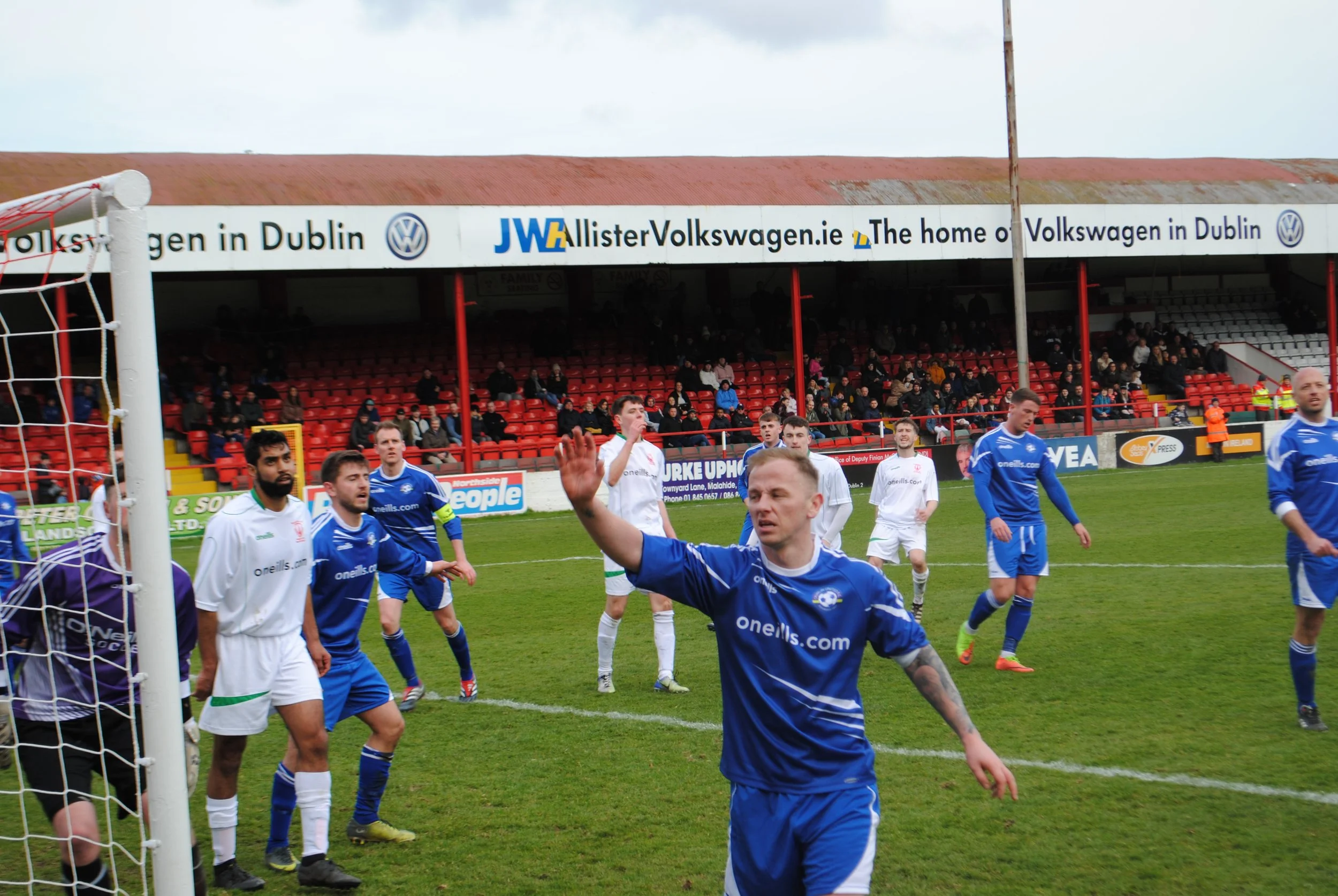 Soccer players on the field near the goalpost, some celebrating, with a stadium and spectators in the background.