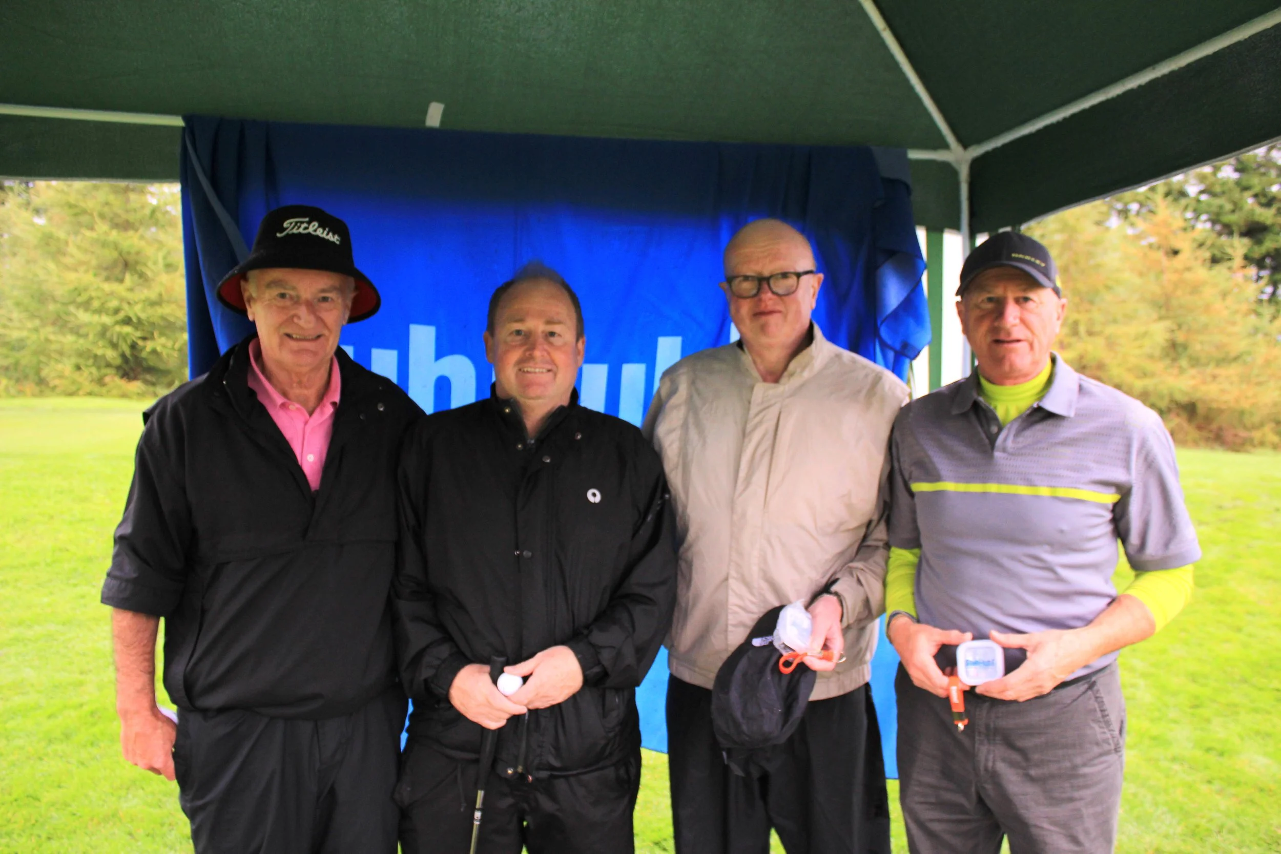 Four men standing together outdoors under a green canopy, with a blue backdrop behind them, likely at a golf event. They are dressed in casual golf or outdoor clothing, and three are holding or wearing golfing accessories.