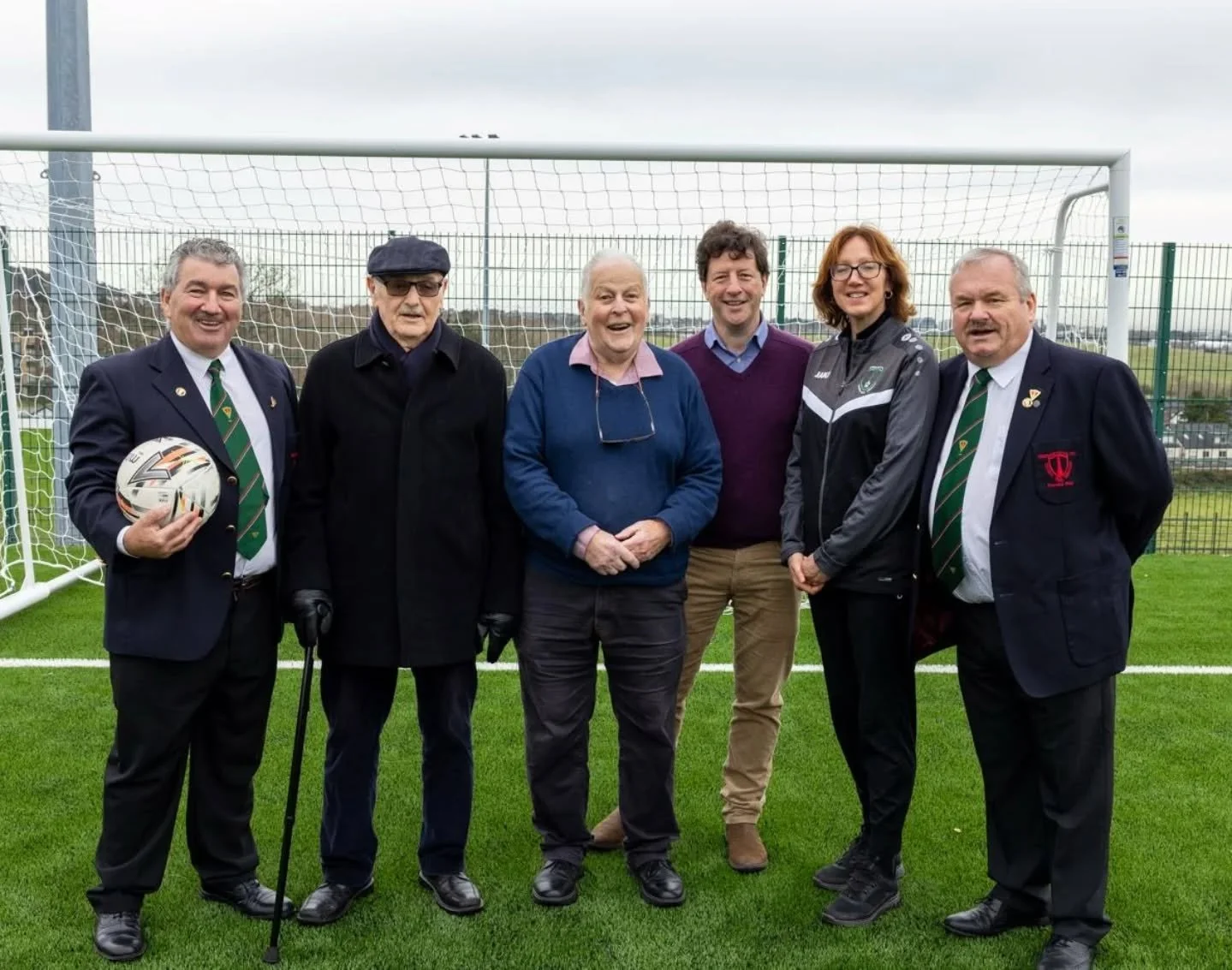 Six people standing on a soccer field in front of a goalpost, smiling for the photo, with three men in suits and three in casual or sportswear, holding a soccer ball and wearing medals.