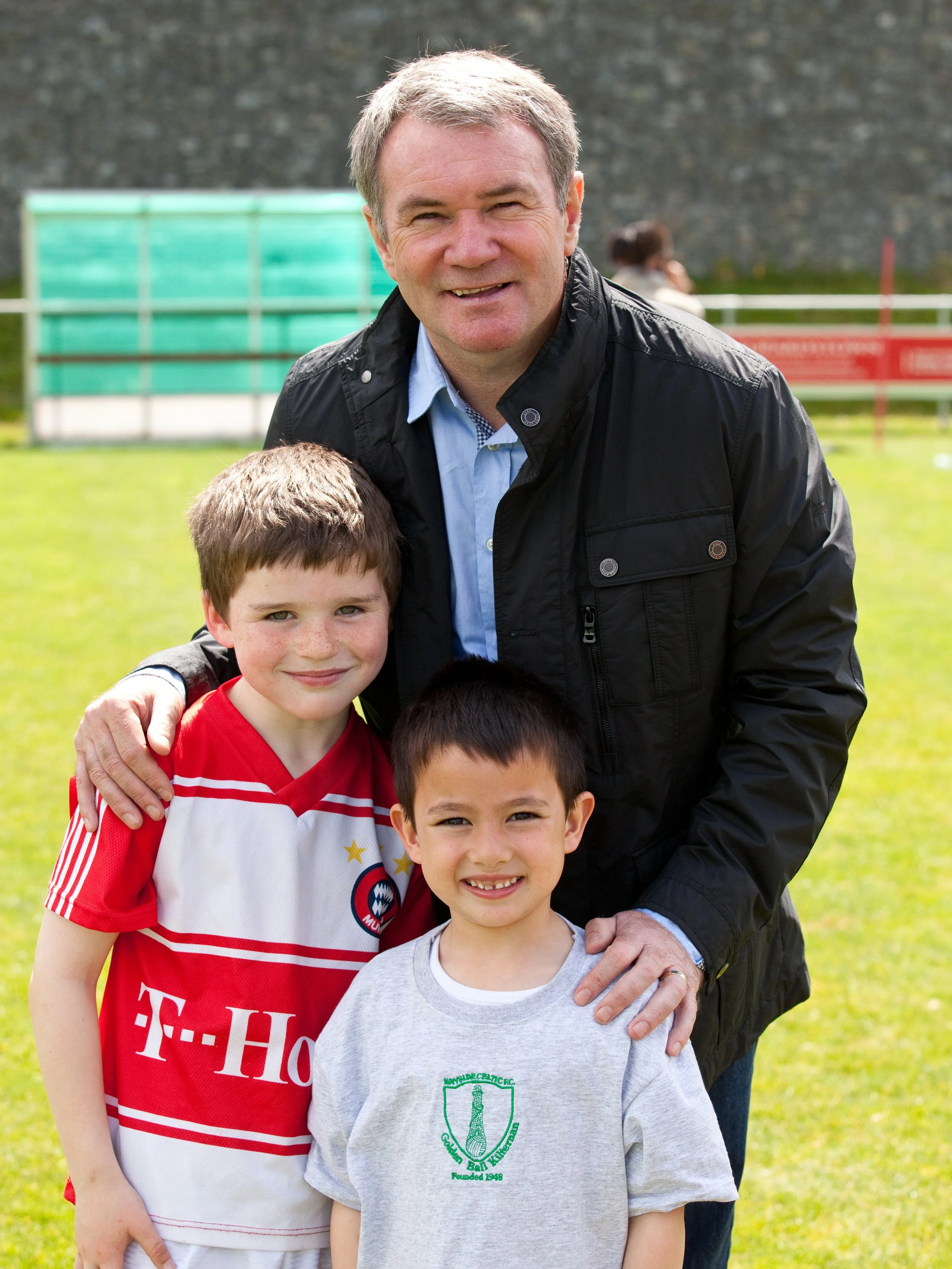 A man in a black jacket and light blue shirt standing outdoors with three young boys on a grassy field, with a stone wall and some spectators in the background.