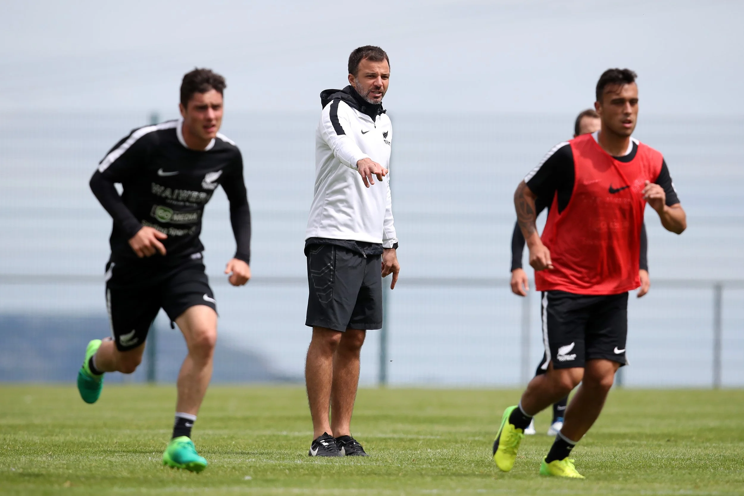 A group of athletes jogging on a grassy field with a coach or trainer giving instructions in the center, during an outdoor training session.