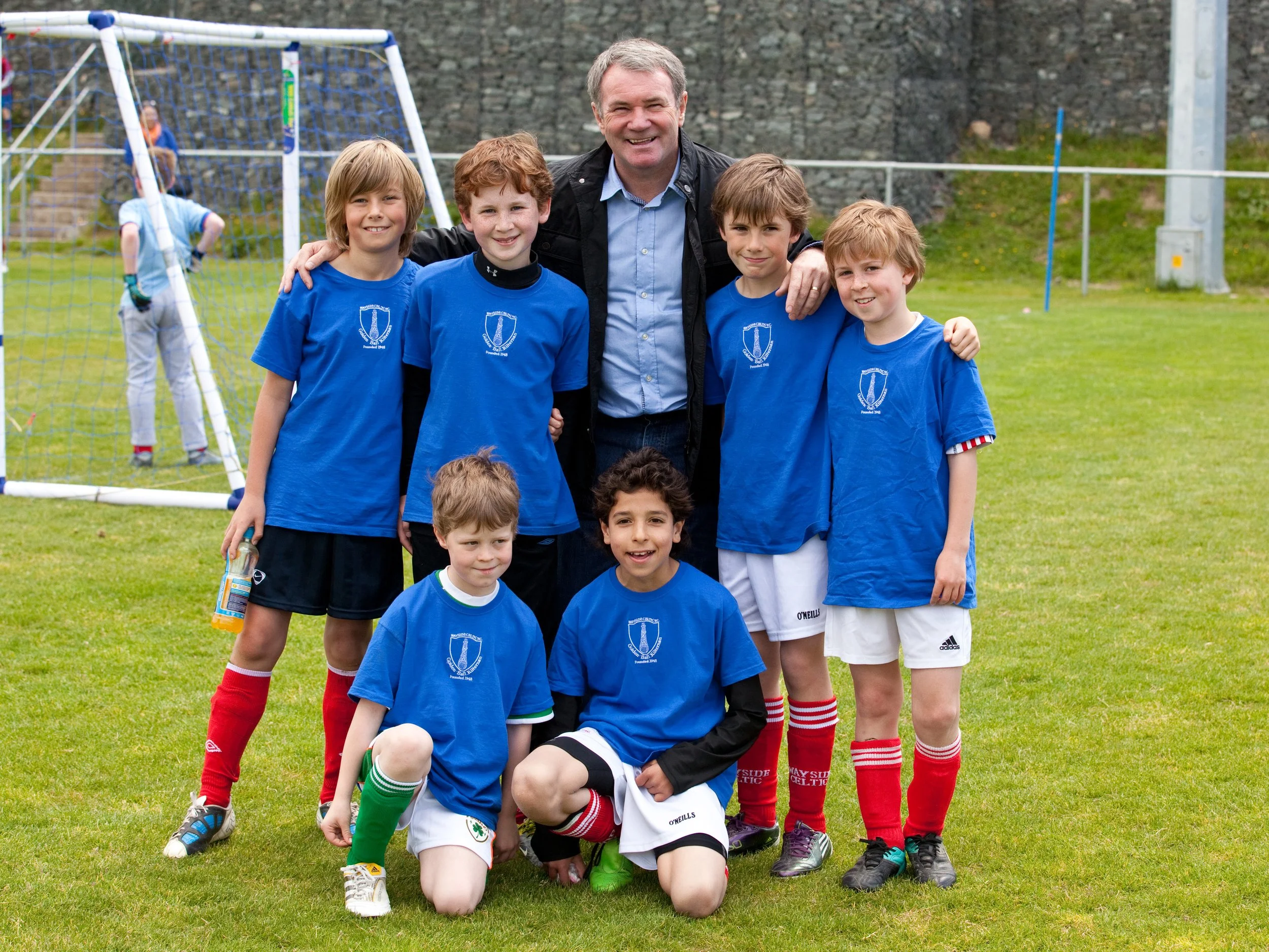 A group of young boys in blue soccer jerseys and red socks, along with a man, pose for a team picture on a soccer field, with a soccer goal and other children in the background.