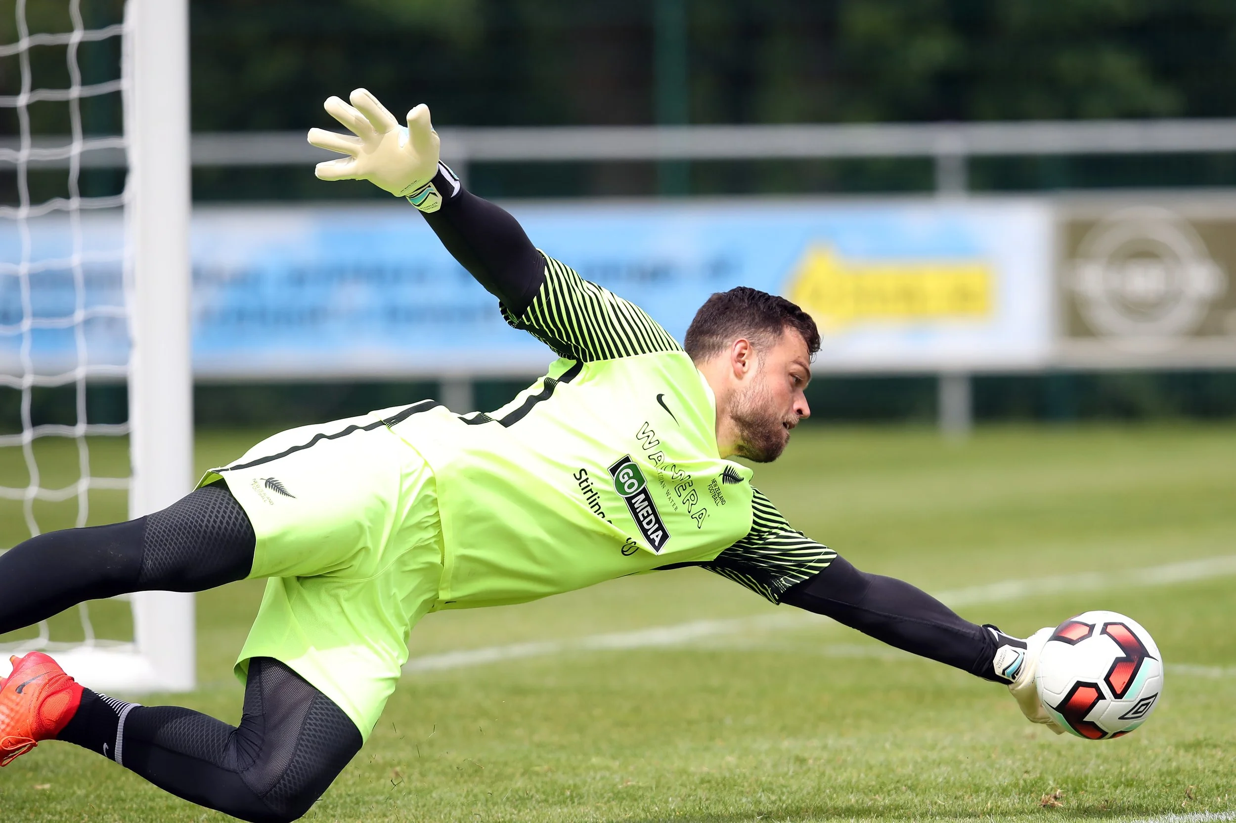 A male soccer goalkeeper in a green uniform diving to save a soccer ball on a grassy field.