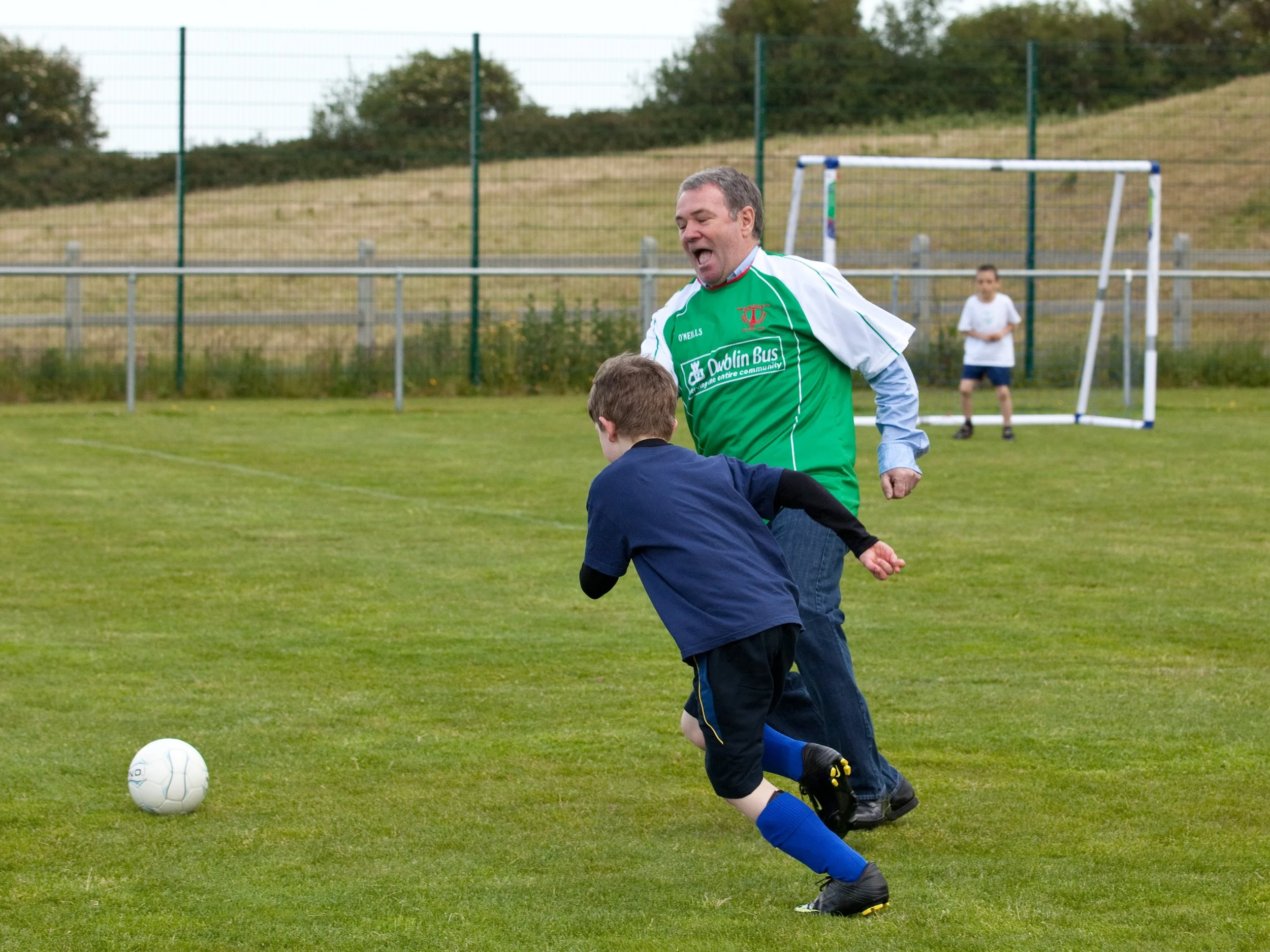 A man and a young boy playing soccer on a grassy field. The man is wearing a green sports jersey and appears to be celebrating or cheering. There is a soccer ball on the ground, and a goalpost with a goalkeeper in the background. The scene is outdoor