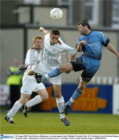Two soccer players in mid-air battle for the ball during a match, with a third player in the background on the field.