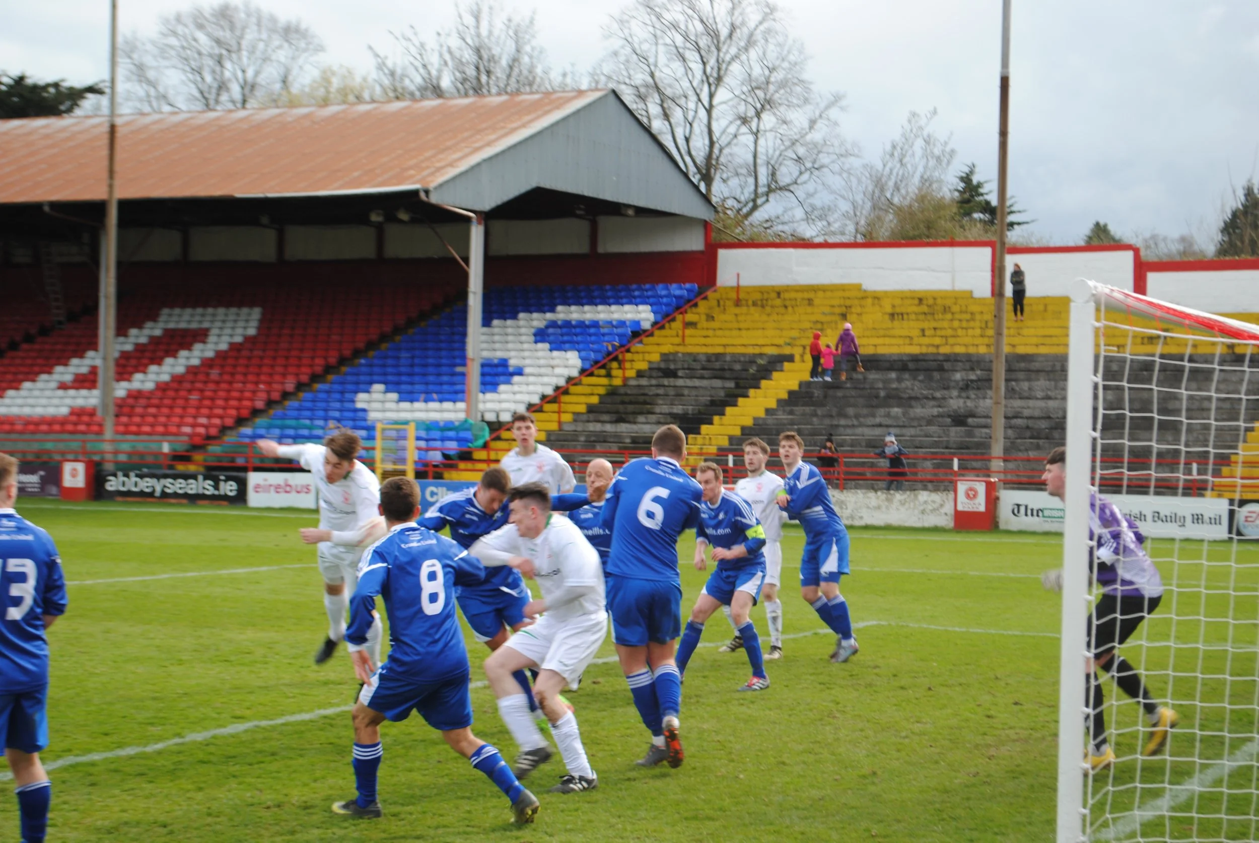 Soccer players in blue and white uniforms competing for the ball during a match on a green field. In the background, empty bleachers with a painted pattern and trees.