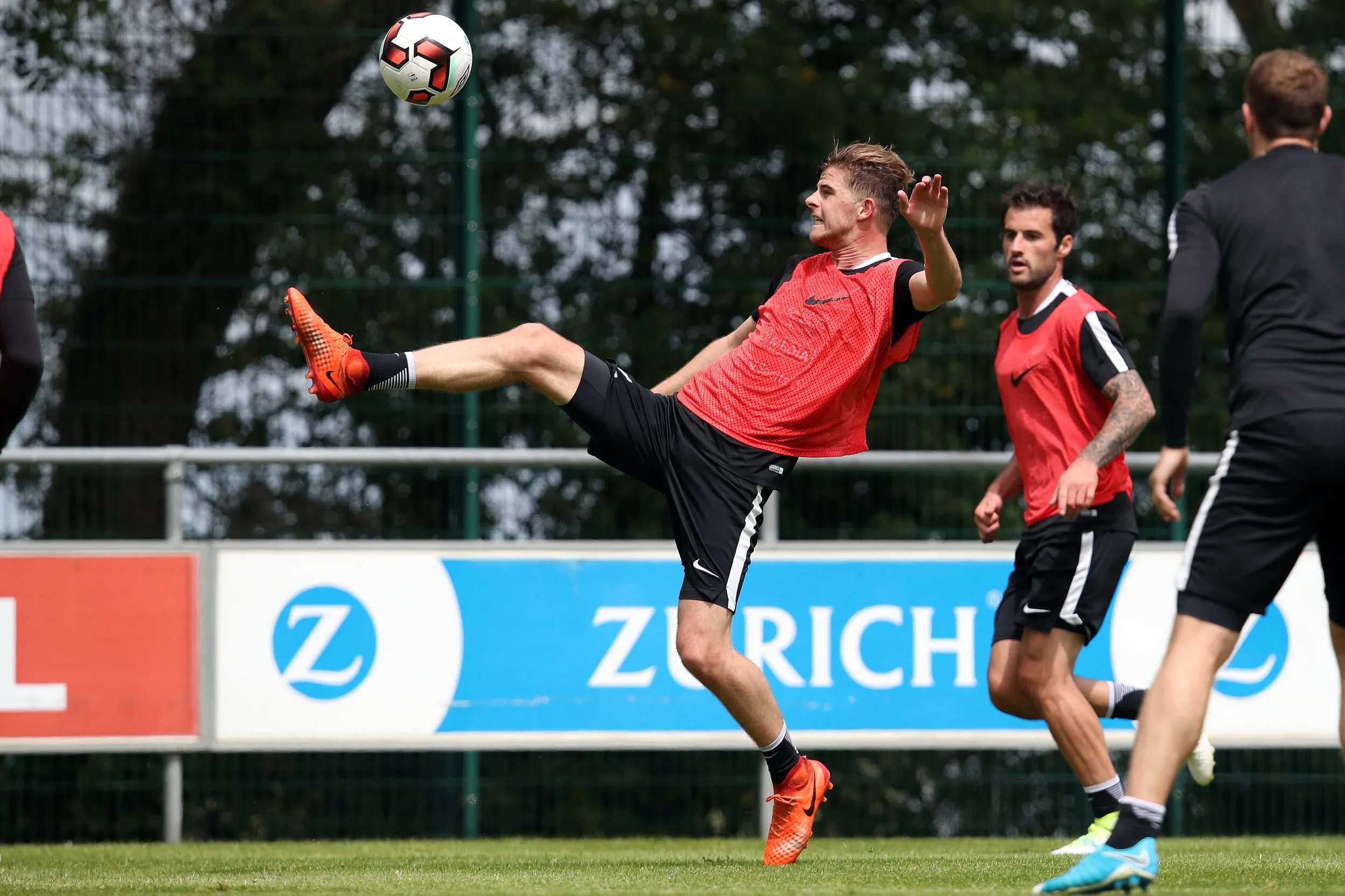 A soccer player in red and black uniform making a high kick for the ball during a game on a grassy field.