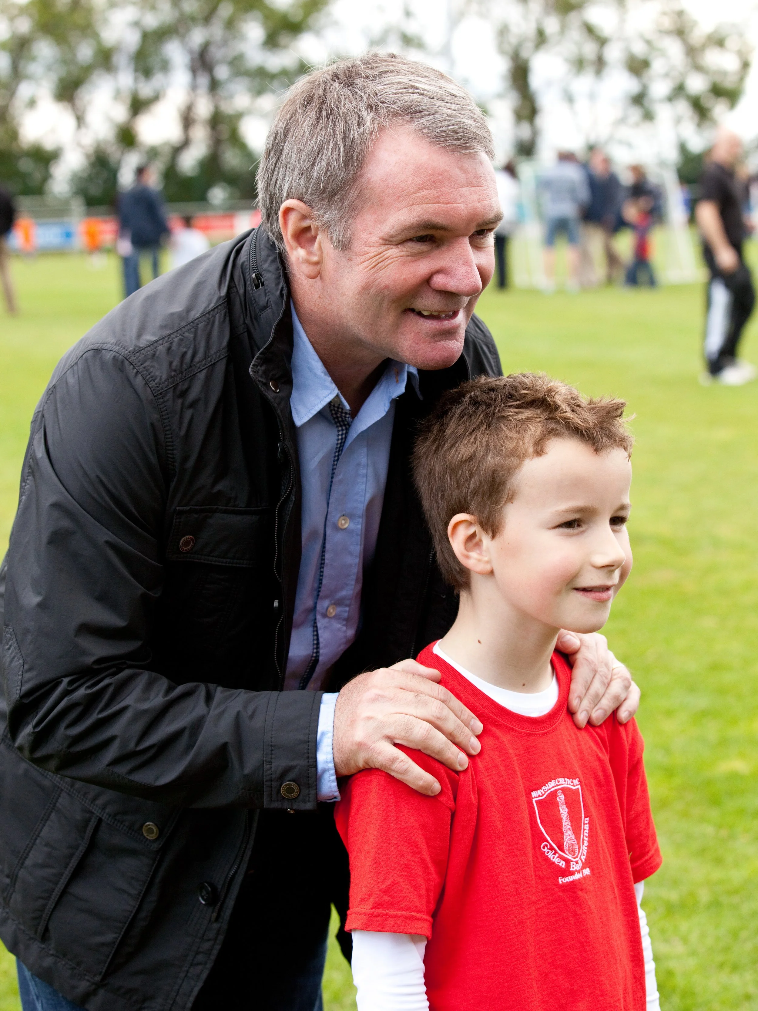 An adult man with gray hair and a young boy with brown hair smiling at a sports event on a grassy field, with other people in the background.