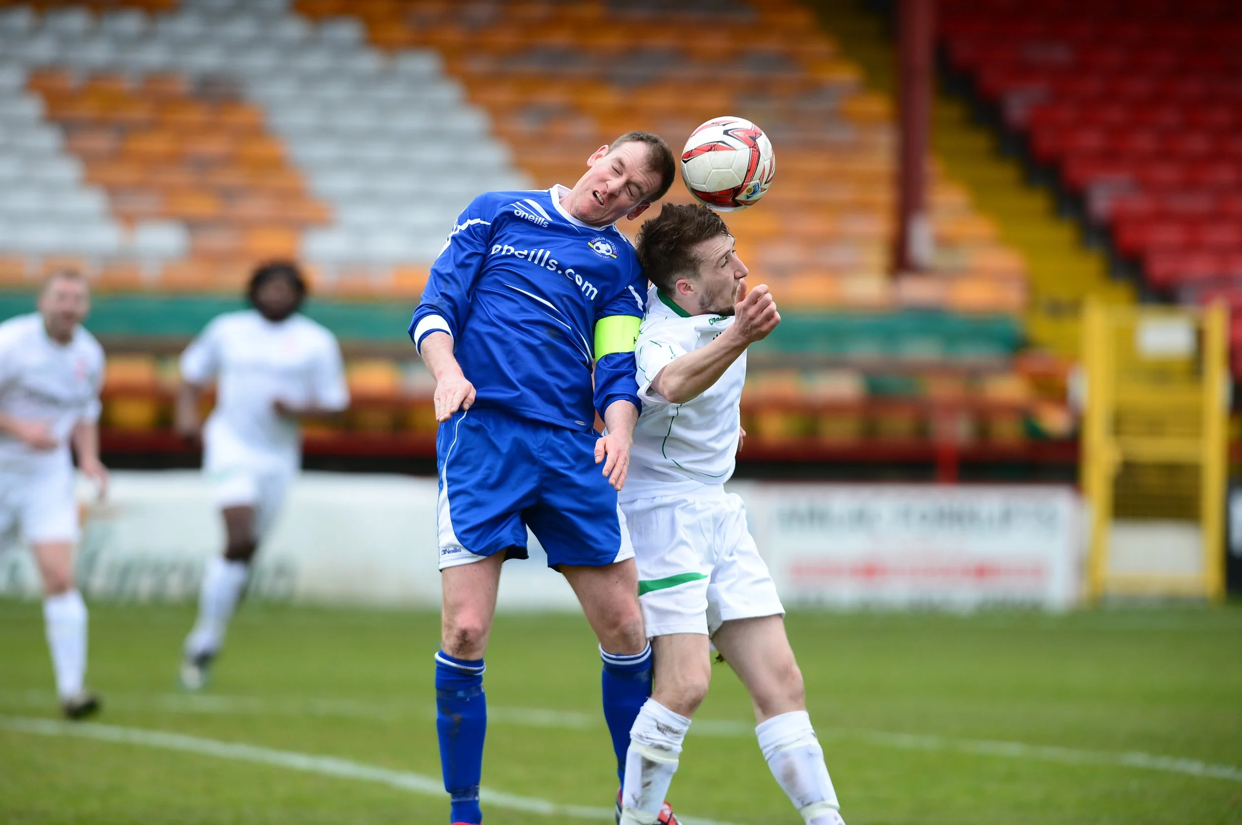 Two soccer players jumping to head the ball during a game, with spectators in the background.
