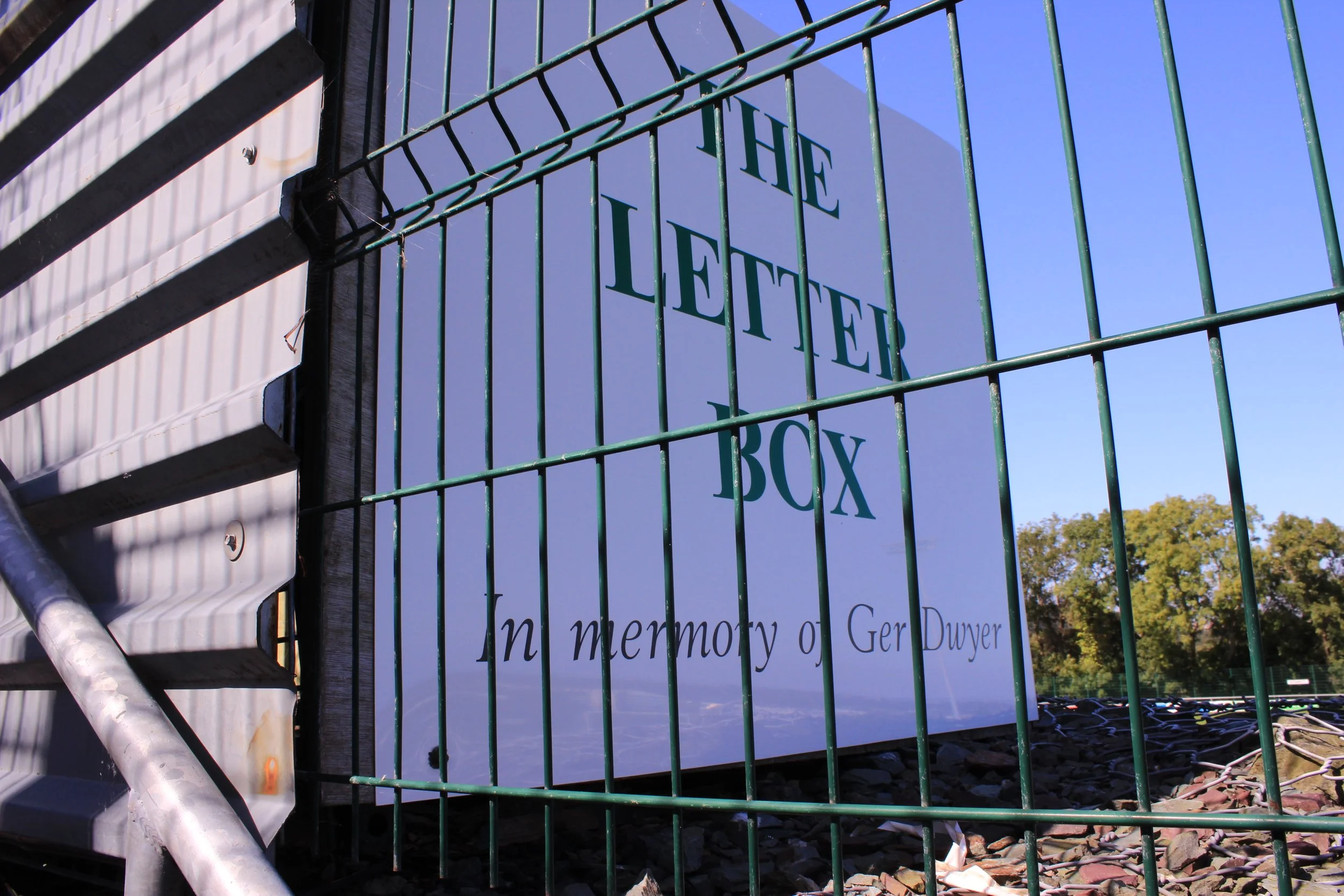 Close-up of a green metal fence with a sign attached to it, with trees and a clear blue sky in the background.